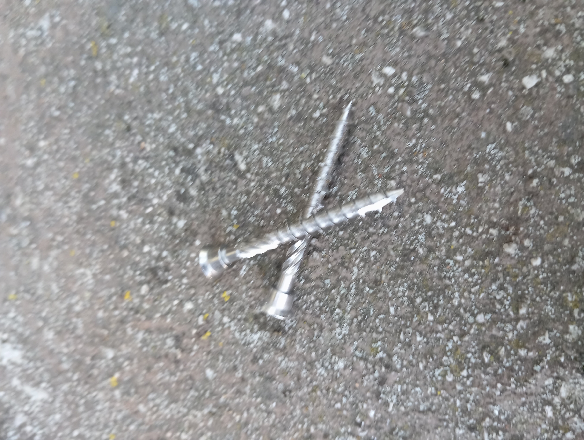 Two silver metal screws are lying on a rough, grey concrete surface. The screws are positioned at an angle, forming an 'X' shape. One screw is positioned horizontally, with its head to the left and its pointed end towards the right. The other screw is diagonally placed across the first, with its head at the top and its pointed end extending downwards and to the left. The concrete surface appears textured and speckled with white and yellow particles. The image is taken from a slightly elevated perspective. The specific location in Baden, Austria cannot be determined from the provided image.