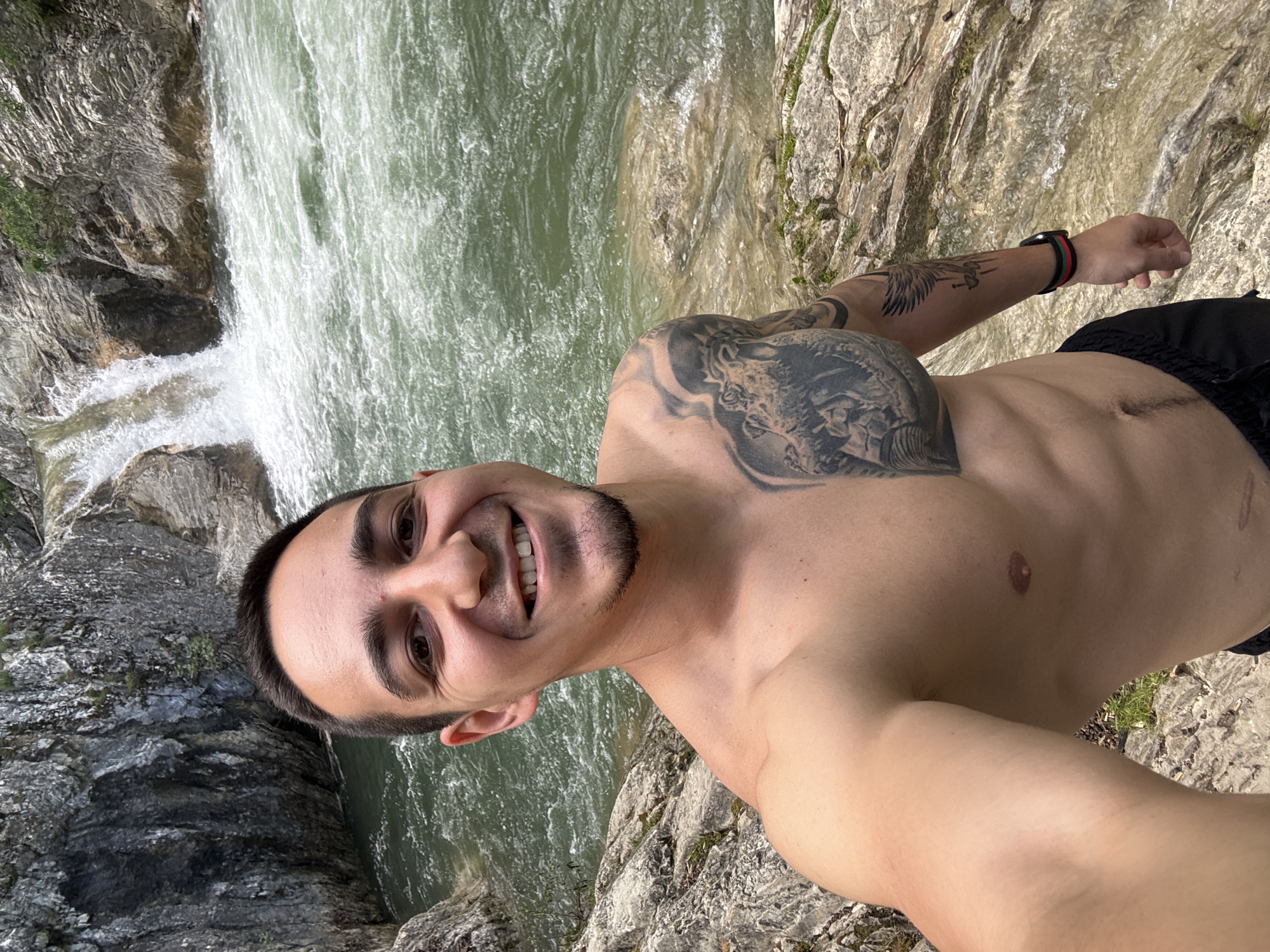 A shirtless man smiles directly at the camera, holding it at arm's length. He stands on a rocky outcrop adjacent to a waterfall and a rushing river. The man has dark hair, a trimmed beard, and tattoos covering his chest and upper arm. He wears a wristband on his left wrist and black shorts. The scene appears to be located outdoors, possibly near Medven, Bulgaria, given the implied location. The focus is on the man and the natural backdrop of water and rock.