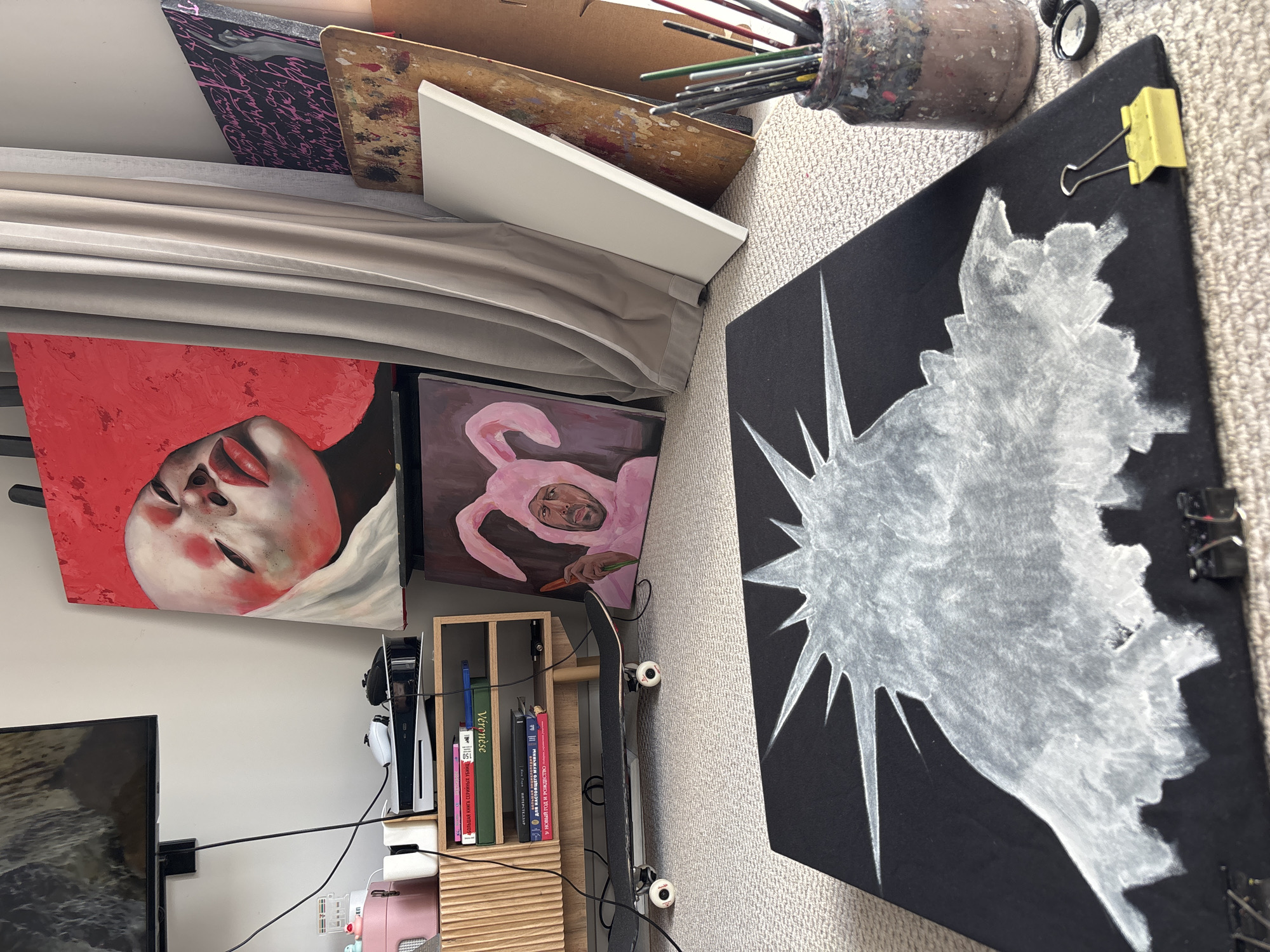 The image shows a cluttered artistic workspace or storage area. Several canvases are visible, including one with a portrait of a person with a red background, another with a person in a pink bunny suit holding a carrot, and another with a white abstract or figurative design on a black background. There is also a stack of canvases and painting supplies such as a jar of paintbrushes.
Below the canvases, there's a small shelf with books and a PlayStation 5 console. A skateboard leans against the shelf. A TV screen can be seen on the left side of the image.
The text on the book spines in Russian appears to read "For a Real Man," "New Year and Christmas," "Veronese," and "Large Book of Serial Killers."
There is no indication of the city of Krasnoyarsk, Russia in the image.