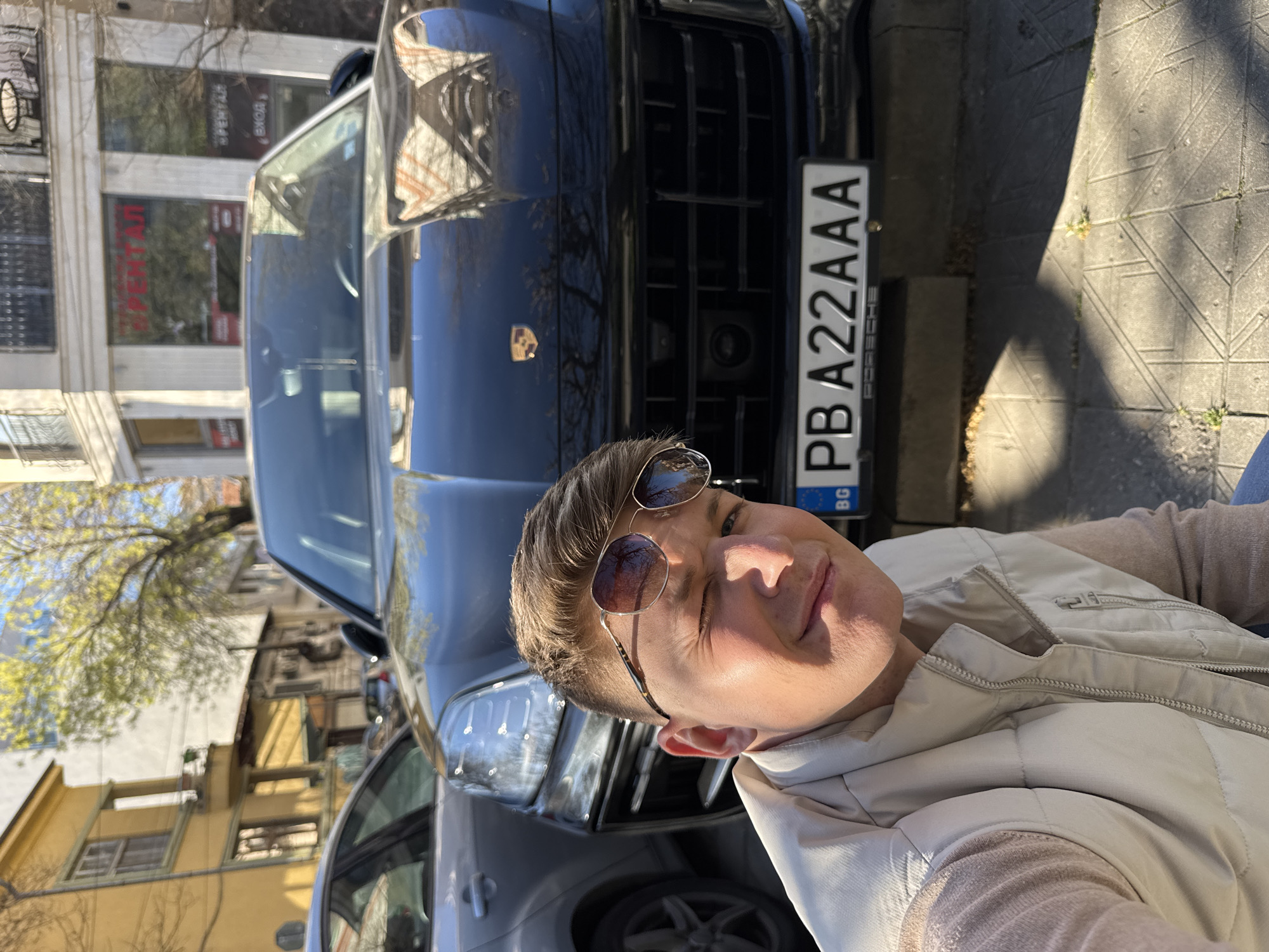 The image shows a person posing in front of a dark-colored Porsche SUV in Plovdiv, Bulgaria. The license plate number is PBA22AAA. The person is wearing sunglasses and a light-colored vest. The background includes buildings, trees with budding leaves, and a sidewalk. The overall environment suggests an urban street setting. The person is likely taking a selfie or having someone take a picture of them with the car.