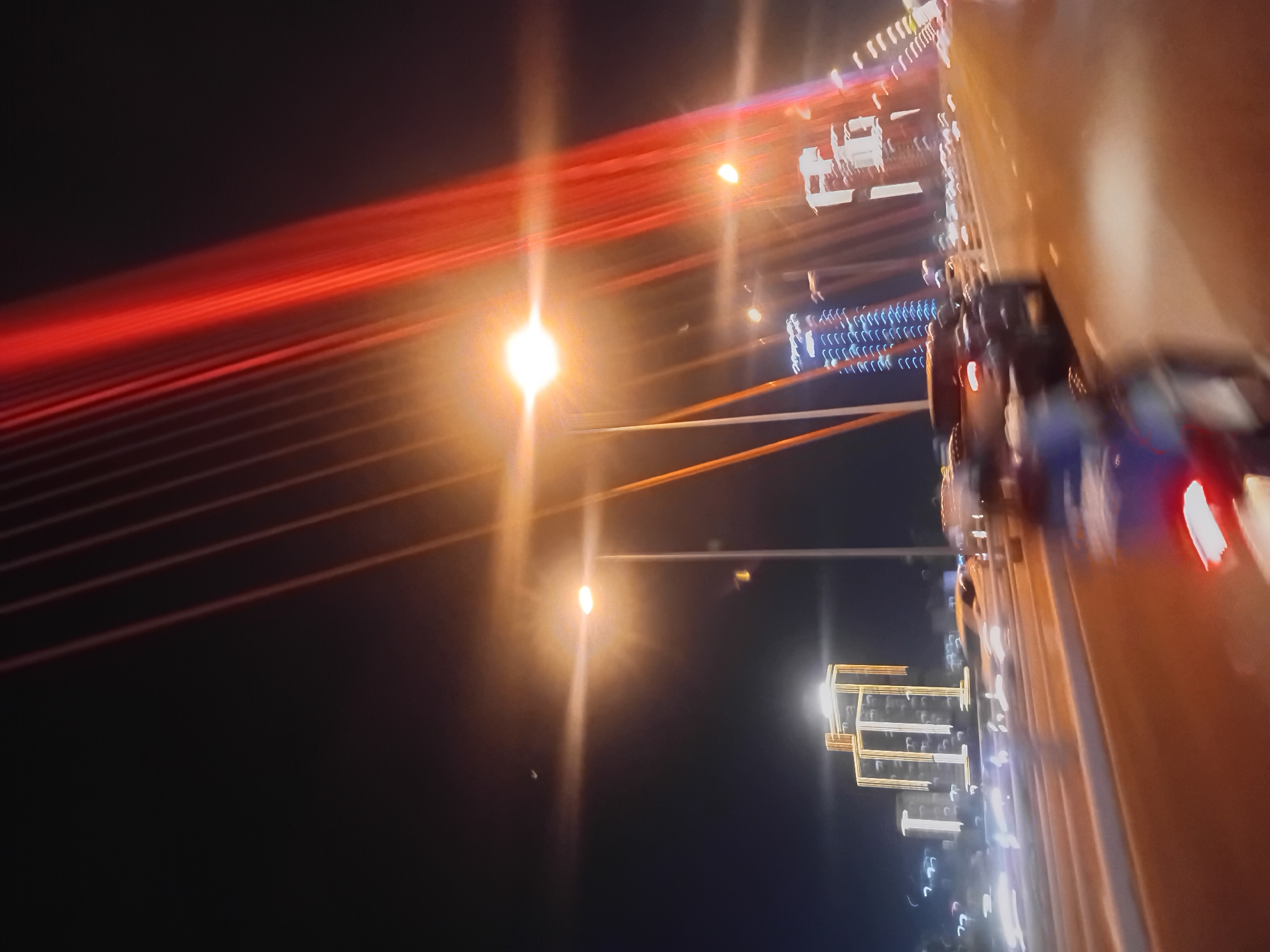 The image is a nighttime long-exposure photograph taken from a moving vehicle on a bridge, likely in Da Nang, Vietnam, given the context. The primary subjects are the illuminated cables of a suspension bridge, which appear as thick, vibrant red streaks stretching diagonally across the frame from the left to the right. These red lights contrast with the dark night sky.
In the mid-ground, a series of bright white and yellow lights are visible, possibly from buildings or decorative lighting along the bridge or on the distant shore. Further to the right, there's a blur of lights from other vehicles, including what appears to be the taillight of a red vehicle in the foreground, also rendered as a streak due to the motion. The right side of the image is dominated by the blurred orange-brown surface of the vehicle the photo was taken from, possibly a truck bed or the side of a van, with its own reflections and lights.
The activity depicted is the movement of traffic across the bridge at night, with the long exposure capturing the dynamic streaks of light from the bridge's structure and other vehicles. The time of day is clearly night, and the weather appears clear, as evidenced by the sharp, bright lights. There are no discernible people in the image, only the effects of light and motion. No visible text is present. The overall impression is one of urban dynamism and the beauty of artificial light at night, enhanced by the motion blur.