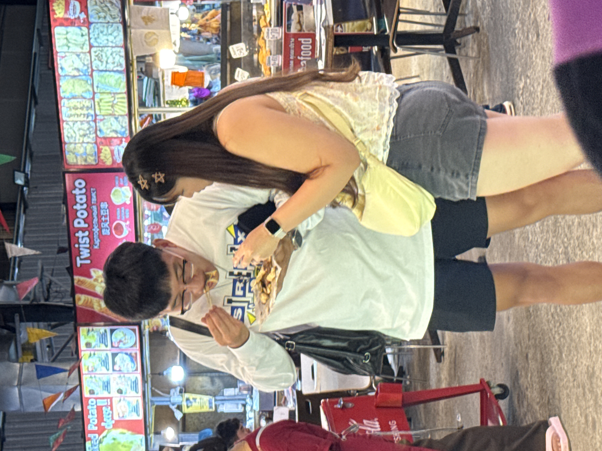 The image depicts a couple at an outdoor food stall, likely in Karon, Thailand. The man in the foreground is wearing glasses and a white long-sleeved shirt, and is in the process of eating fries from a paper container with chopsticks. He appears to be in his early 20s. The woman behind him is wearing a yellow floral top and denim shorts. Her hair is long and dark, adorned with star-shaped clips. She is leaning over, looking at the food. A bright yellow bag is slung over her shoulder.
The food stall has a prominent sign advertising "Twist Potato" with text in English, Russian, and Chinese characters. There are also images of french fries and other fried items on display. Another sign reads "food." The overall setting suggests a vibrant night market or a bustling street food area. Colorful triangular flags are strung up in the background. The lighting appears to be artificial, indicating it's likely evening or nighttime. The overall mood seems casual and enjoyable, as the couple shares a snack.