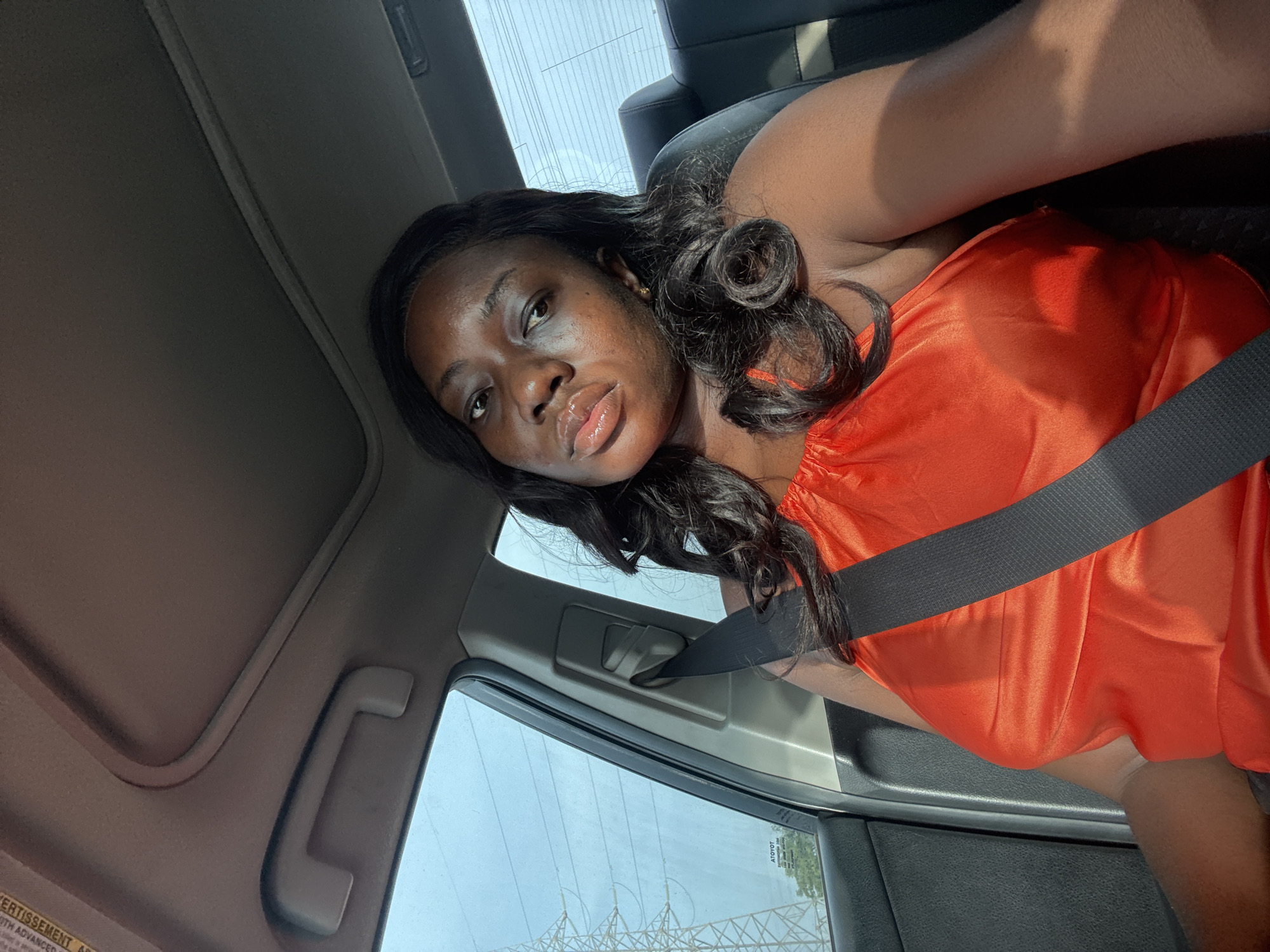 A medium shot depicts a dark-skinned female with curled black hair. She is wearing an orange, silky garment and is restrained by a black seatbelt. The woman is positioned inside a vehicle, potentially in the driver's seat. The interior includes elements such as the headliner, window, and door frame. Through the window, a view of electrical towers and power lines is discernible. The location is identified as Kuchingoro, Nigeria.