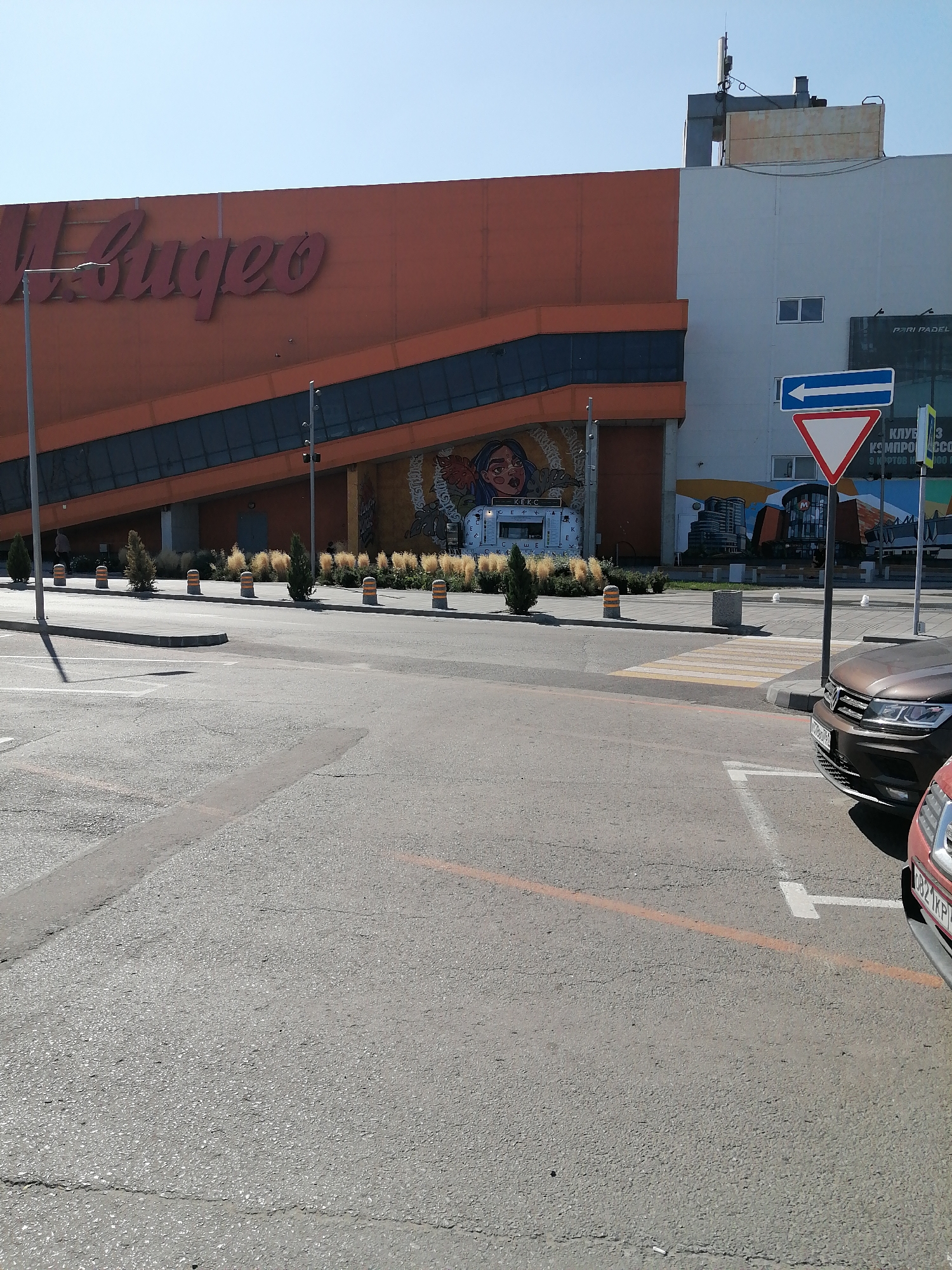 The image depicts an outdoor scene in front of a large building with an orange facade featuring red lettering that reads "М.Видео". The building appears to be a commercial establishment, possibly a electronics store. The facade has a dark blue diagonal band running across it, and further down, there is mural art depicting a stylized face and city buildings.
To the right of the building, a white structure with windows is visible. A street sign with a blue arrow pointing left and a red triangular warning sign is mounted on a pole. Behind the warning sign, there are Cyrillic characters, some of which are legible and indicate "КЛУБ" and "КЭШИР".
In the foreground, there is a paved parking lot with white parking lines. A brown car is parked in the lot, and the front of a red car is also visible. There are a few small trees and decorative bollards lined up along a sidewalk in front of the building. The sky is clear and blue.
Based on the context provided by the text on the building ("М.Видео") and the street signage (Cyrillic script), this location is likely in Russia. The presence of the commercial chain "M.Video" and the visible Cyrillic text strongly suggests a Russian setting. The specific mention of Rostov-on-Don, Russia, points to this particular city as the location.
