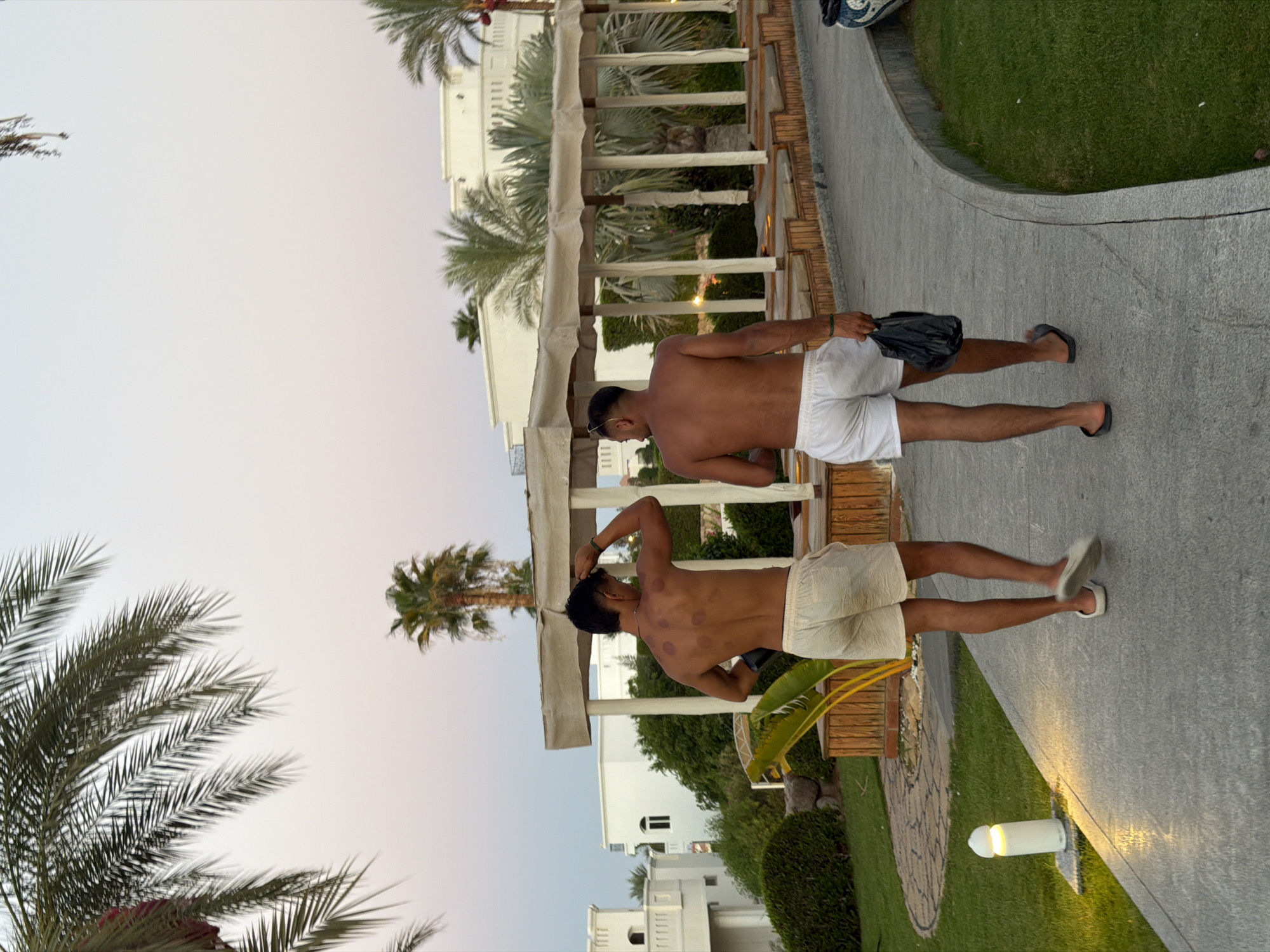 Two men, shirtless and wearing shorts, walk away from the camera on a paved path in Sharm El Sheikh, Egypt. The man on the left has several circular bruises on his back, consistent with cupping therapy. He is wearing light-colored shorts, and the man on the right is wearing white shorts. Both men are wearing sandals. The path is bordered by green grass and palm trees. In the background, there are white buildings with arched windows and a covered outdoor seating area with a canopy. The sky is a soft pink and orange hue, suggesting it is either dawn or dusk. A white, mushroom-shaped outdoor lamp is visible to the left of the path, illuminated and casting a warm glow on the grass.