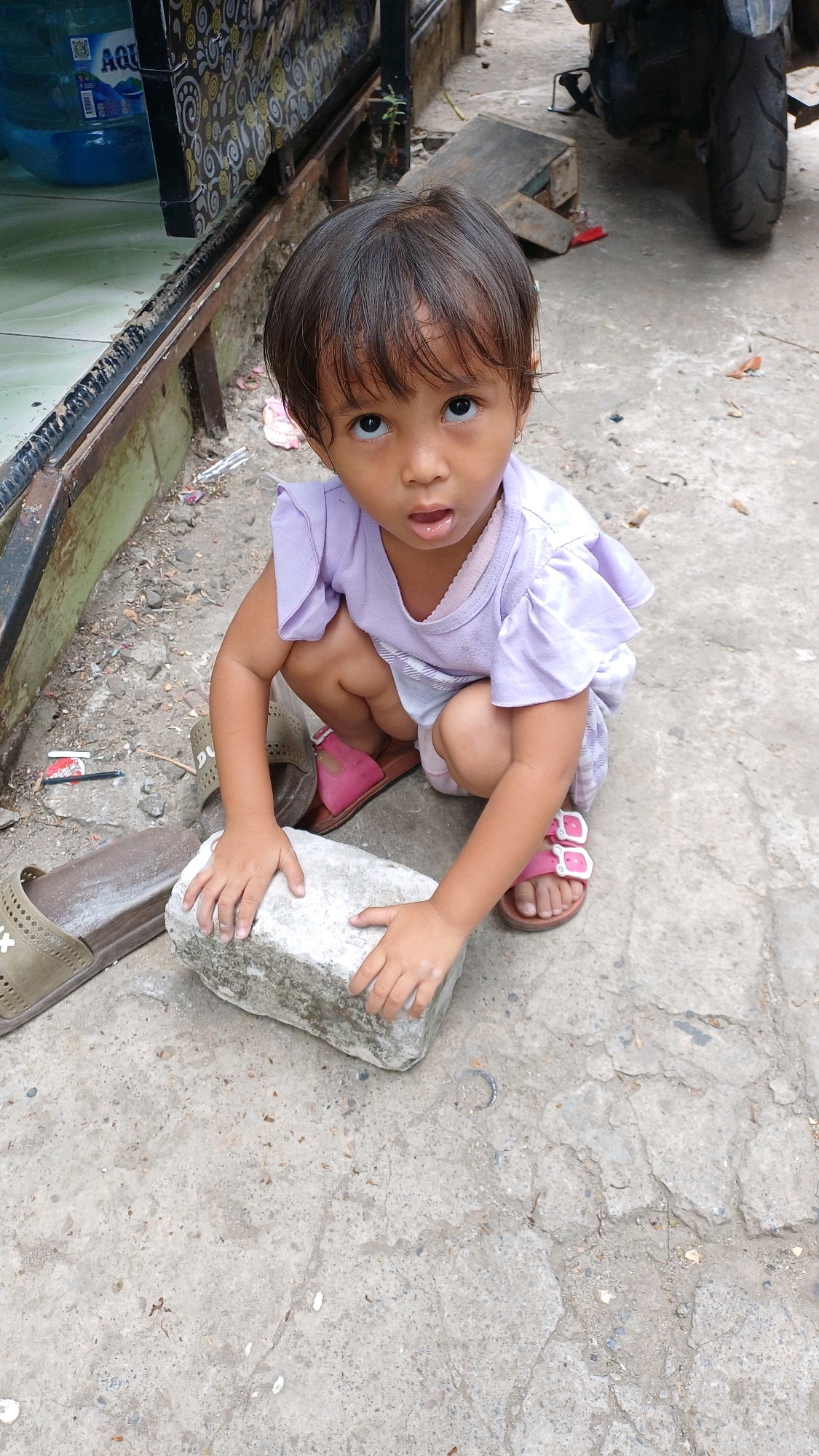 A young child, possibly a girl based on her attire, is crouching on a concrete surface in Gunung Putri, Indonesia. She is wearing a light purple top with ruffled sleeves and white bottoms. Her feet are adorned with pink sandals. In front of her, she is interacting with a rectangular grey object, possibly a piece of concrete or a brick, with both hands. To her left, a brown sandal is lying on the ground, and to her right, a pink sandal is visible. The environment appears to be outdoors, with a textured concrete ground and some debris scattered around. In the background, a portion of a structure with decorative panels and a large blue water jug are visible. A motorcycle is also partially visible in the background to the right. The lighting suggests it is daytime.
