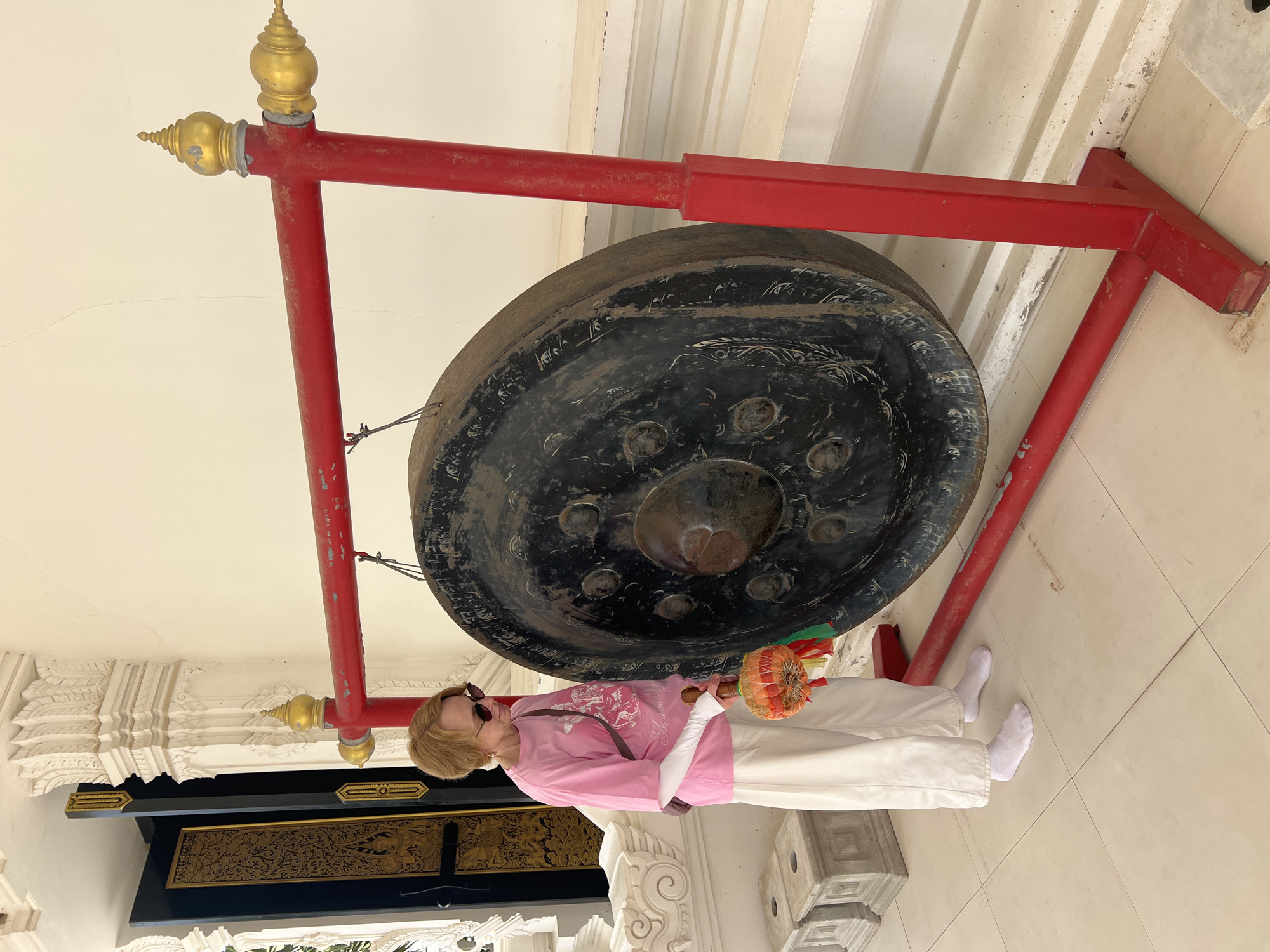 A woman stands beside a large, black gong hanging from a red metal frame. She is wearing a pink t-shirt, white pants, white socks, and sunglasses. She holds a gong mallet with an orange and green decorative end. The gong has an intricate design visible on its surface. The scene appears to be outdoors, possibly at a temple or cultural site, given the ornate decorative elements and the presence of the gong, which is often used in spiritual or ceremonial contexts. The background shows white pillars and architectural details, suggesting a tropical or Asian setting. The lighting suggests it is daytime. The woman's expression is not clearly visible due to her sunglasses and the angle. The ground is tiled. There is no visible text in the image.