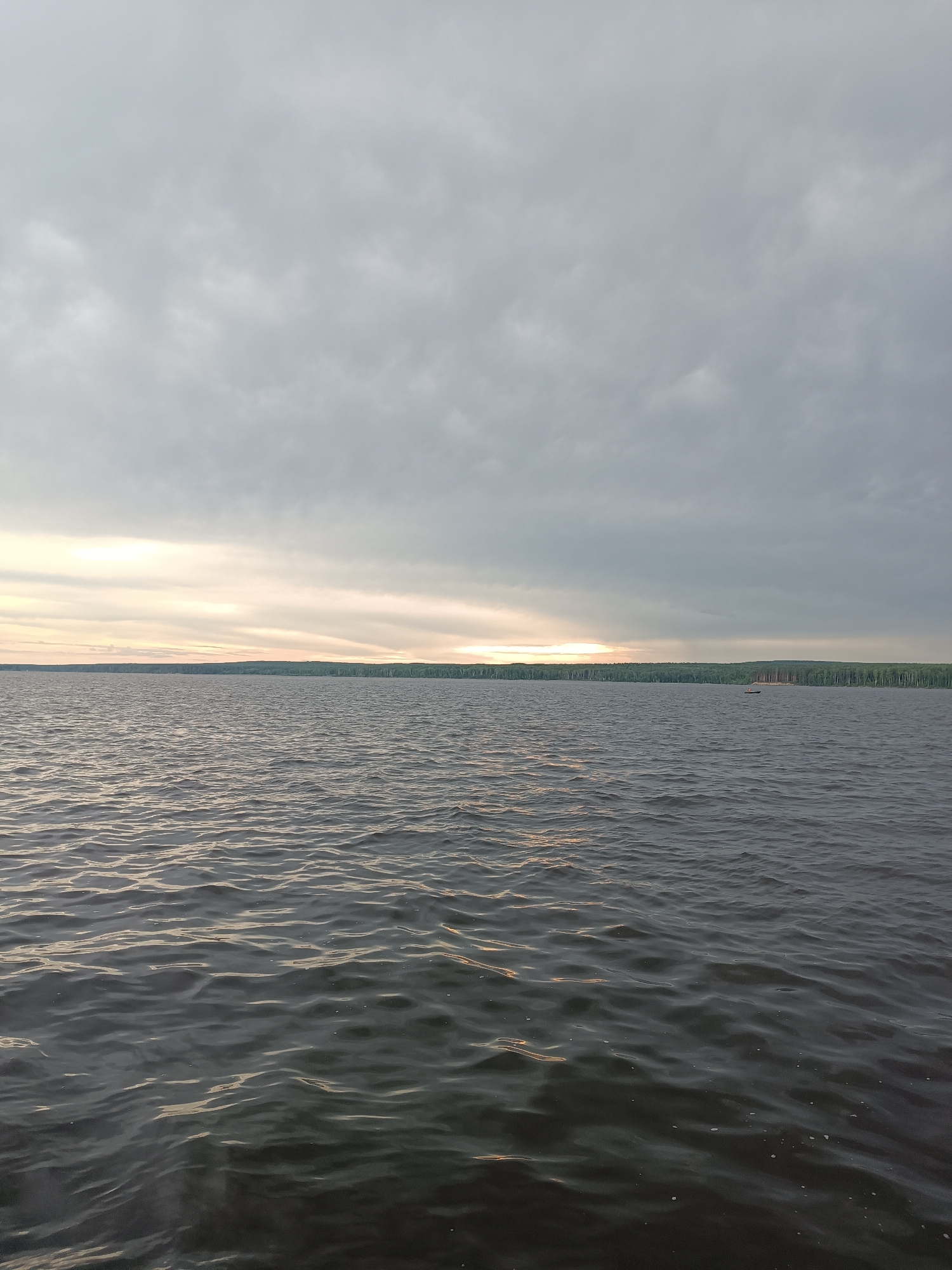 A wide shot captures a body of water under a cloudy sky. The water is dark and rippled, reflecting the muted light of the sky. A distant shoreline is visible, lined with trees and some structures, suggesting it may be near a populated area. The sky is overcast with bands of lighter clouds towards the horizon, indicating either sunrise or sunset. In the middle distance, a small boat is visible on the water. The image appears to have been taken from another boat or the shore. The location is the Kama River in the city of Chaikovsky, Russia.