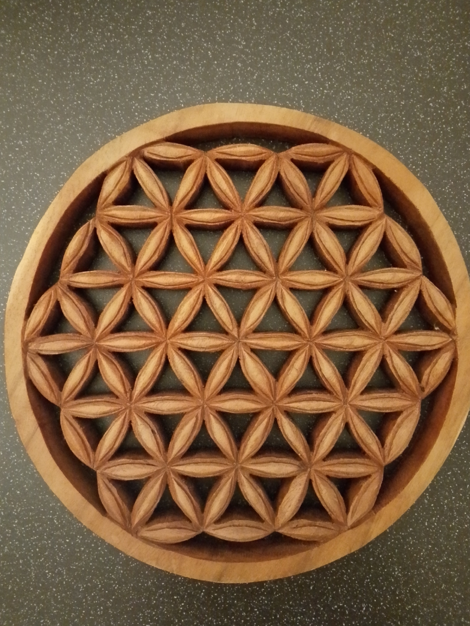 The image shows a wooden carving of the Flower of Life pattern. The carving is set within a circular wooden frame and placed on a dark gray surface with subtle speckles. The pattern is intricate, consisting of overlapping circles that form a series of petal-like shapes. The carving appears to be hand-crafted. There are no people visible. The location is not in 1-Go Maya, Russia, but unknown.