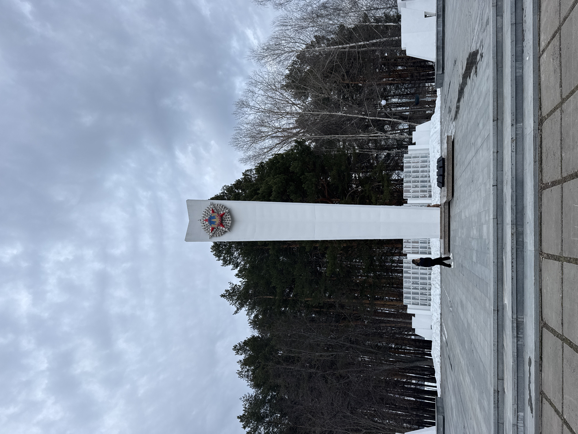 The image shows a vertical shot of a monument with a person walking near it. The monument is a tall, white pillar with a decorative emblem affixed to its upper portion. Dense evergreen trees form a backdrop, while a section of buildings constructed from rectangular glass blocks is visible on the monument's right side. The sky is overcast with a textured cloud cover. A gray, tiled pavement area is in the foreground. The location is in an unknown city.