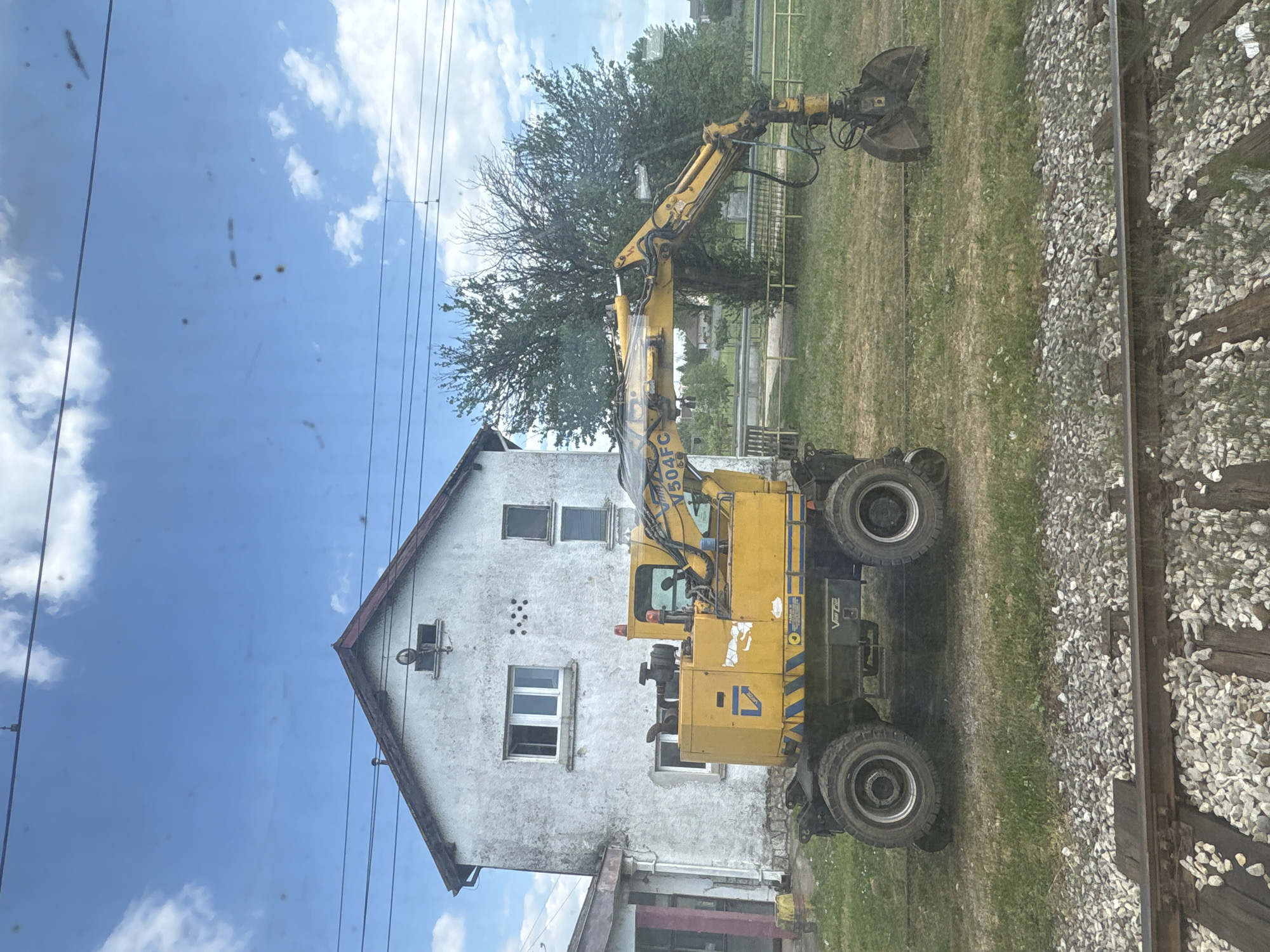 The image, taken in Mahala, Montenegro, presents a daytime scene alongside a railway track under a bright blue sky with scattered white clouds. A prominent yellow wheeled excavator is positioned centrally in the frame, facing right. This industrial vehicle features a long, articulated boom terminating in a grapple or material handling attachment. Large black tires support the excavator, which also displays the text "V504FC" on its boom and "VPTEL" on its side, alongside a distinct blue and yellow striped pattern and a white, stylized animal logo.

To the left of the excavator, a two-story white house with a reddish-brown gabled roof and multiple windows stands. Behind the excavator, a large, green, leafy tree provides a natural backdrop. On the far right, a segment of a railway track is clearly visible, composed of grey ballast stones, dark wooden sleepers, and metal rails. Overhead power lines or cables are strung across the sky. The photograph appears to have been taken through a window, evidenced by reflections and small specks of dirt or dust. The excavator is stationary, indicating it is either parked or paused, with no active work occurring. The scene is calm and showcases a blend of residential, natural, and infrastructural elements.