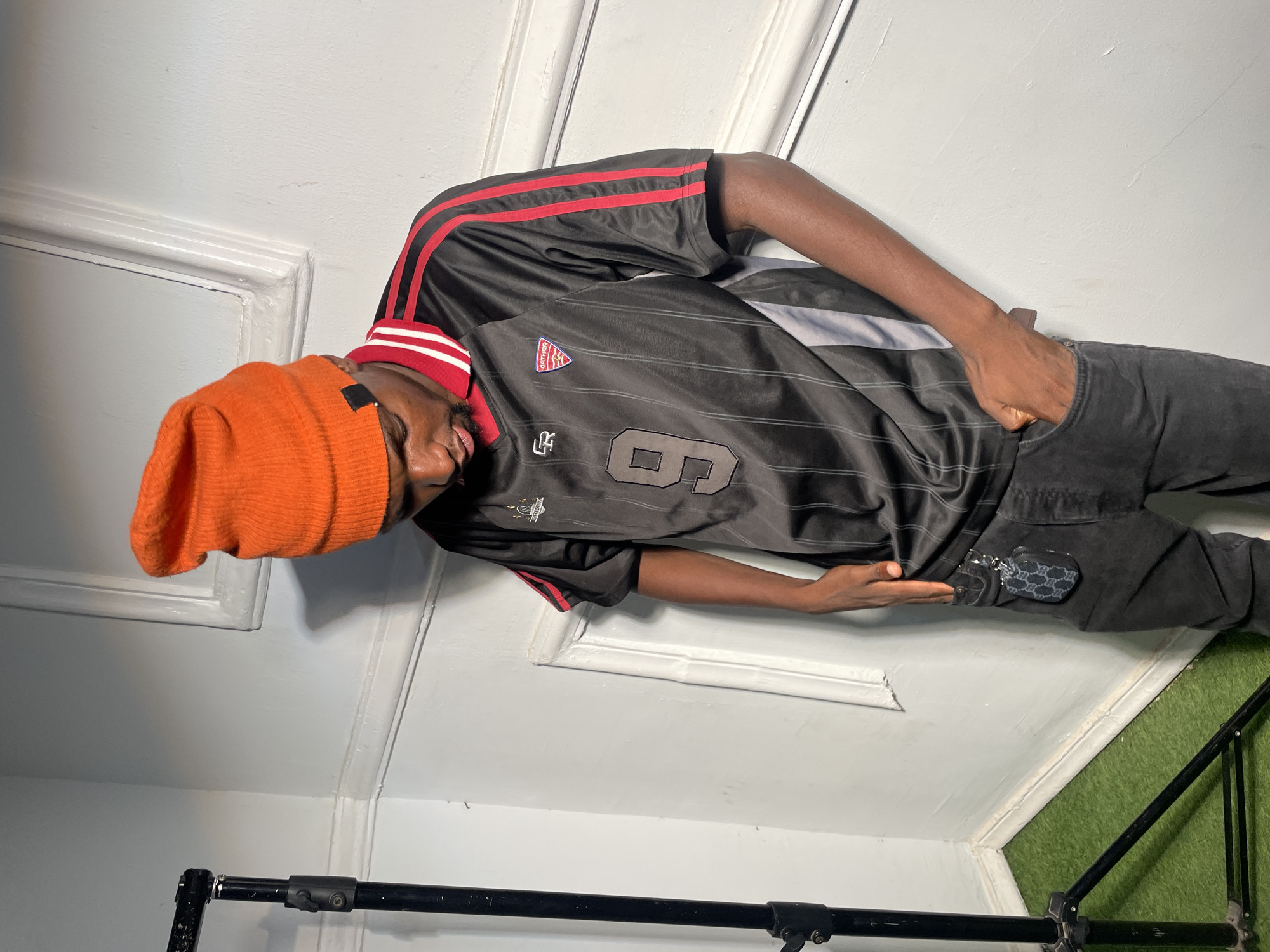 A slender Black man is shown indoors against a white door and wall backdrop. The man is wearing an orange beanie, a black soccer jersey with red stripes on the shoulders, and dark denim pants. He has one hand in his pocket. The number "6" is visible on the jersey. A black tripod stand is visible at the bottom of the frame, and a patch of green, possibly artificial grass, is visible on the lower right. A small dark-colored object, possibly a coin purse or keychain, hangs from his belt loop. There is no specific evidence to place this photo in Barakin Akawo, Nigeria.
