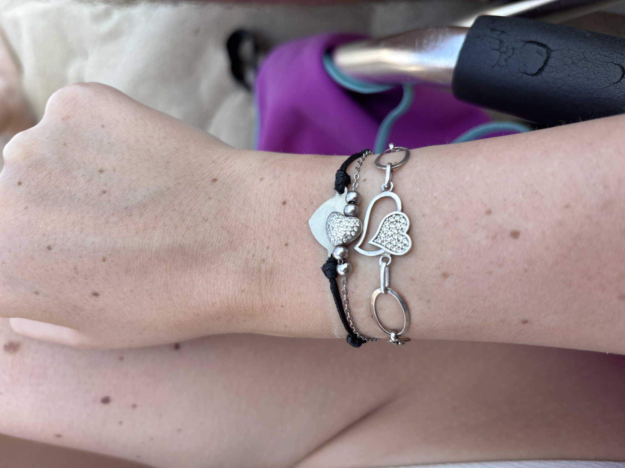 A person's arm, adorned with two silver bracelets, is visible against a background that includes a pink fabric object and part of a black grip. The arm exhibits numerous small, dark moles. The scene is devoid of any urban context, and therefore its location within the city of Kiten, Bulgaria, cannot be determined.