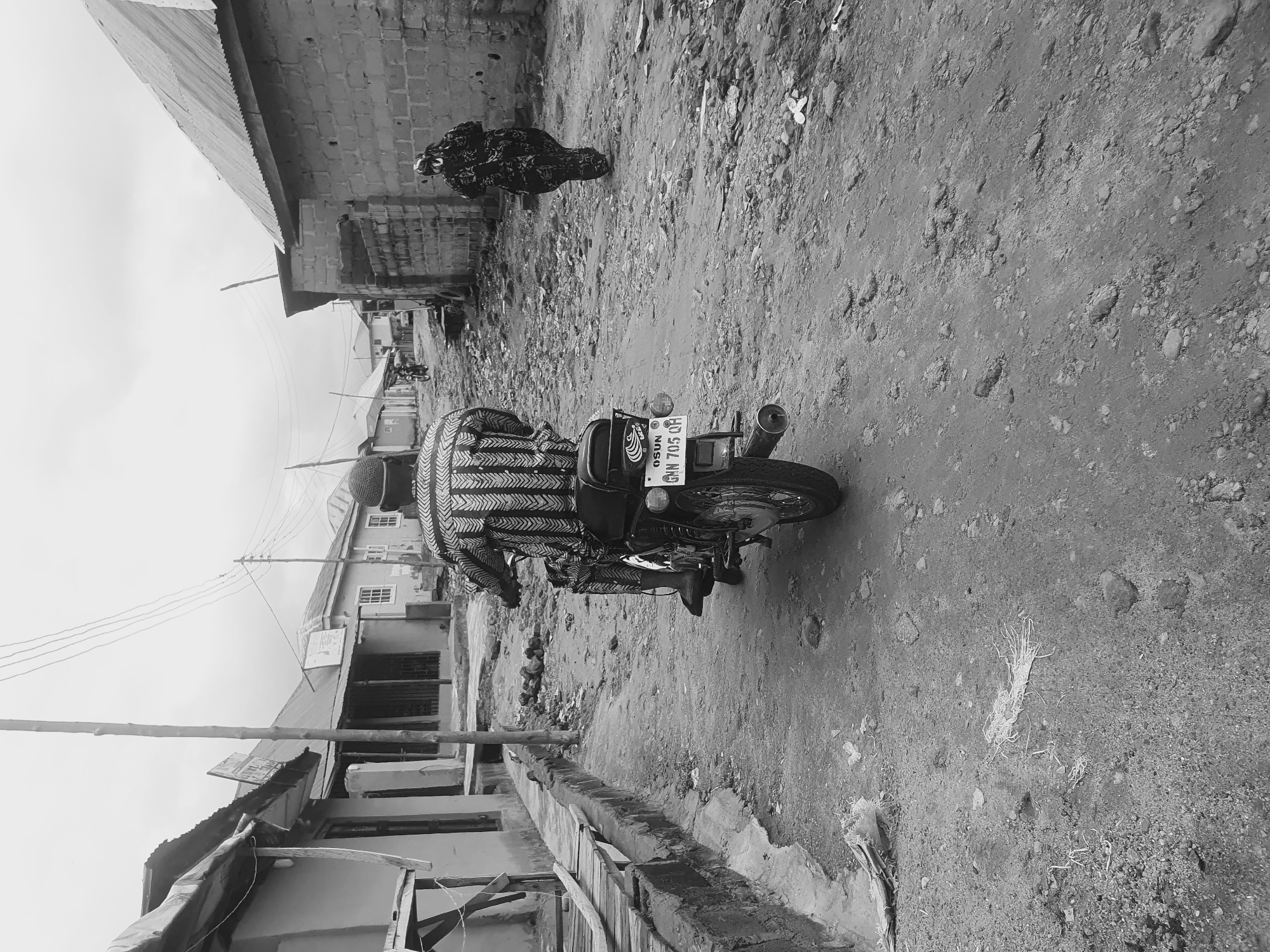 The image shows a street in Lokoja, Nigeria. The scene is in black and white, featuring a man on a motorcycle and another person standing further down the road. The motorcycle's license plate is visible and reads "OSUN GNN 705 QR." The street appears unpaved and lined with buildings of simple construction, indicating a developing or lower-income area. Overhead, electrical wires stretch between poles. The setting suggests a daytime scene, with the lighting and shadows defining the environment.