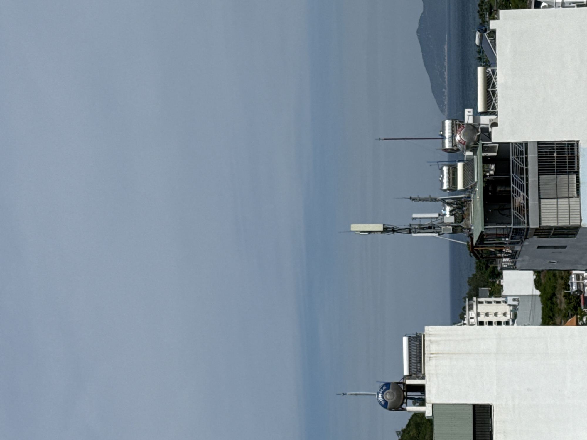 The image captures a cityscape with buildings and the sea in the background. In the foreground, a white building with a green-paneled section is prominent. On top of this building, a blue solar water heater with the word "SONHA" visible is installed. Adjacent to it, a solar panel array is also present. To the right, another white building features multiple solar water heaters and antenna arrays. Further in the background, a multi-story building with visible windows is seen amidst lush green foliage. The sea stretches out towards a distant, mountainous island. The sky is overcast with a pale blue hue. The scene appears to be a rooftop view in a coastal urban area, likely Bac Nha Trang Ward, Vietnam, given the presence of solar water heaters and the coastal landscape. No people are visible in the frame.