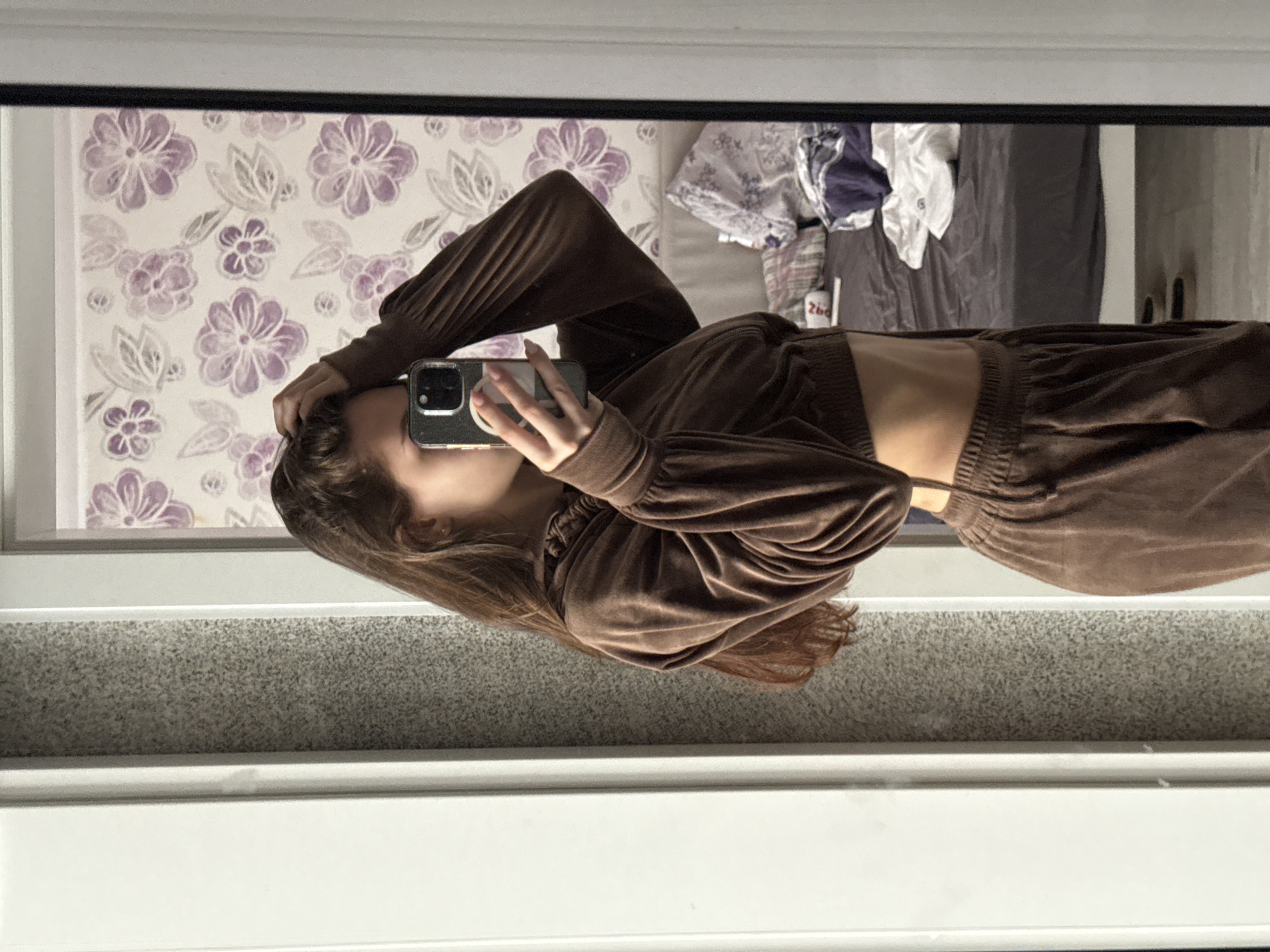 A woman takes a picture of herself in a mirror. The woman has long brown hair and is wearing a brown, two-piece outfit that reveals her midriff. She is holding a phone in her left hand, with her right hand touching her hair. The reflection in the mirror shows a wall with floral wallpaper and a bed with clothes on it. The location is not discernible as Sofia, Bulgaria, based on the provided image.