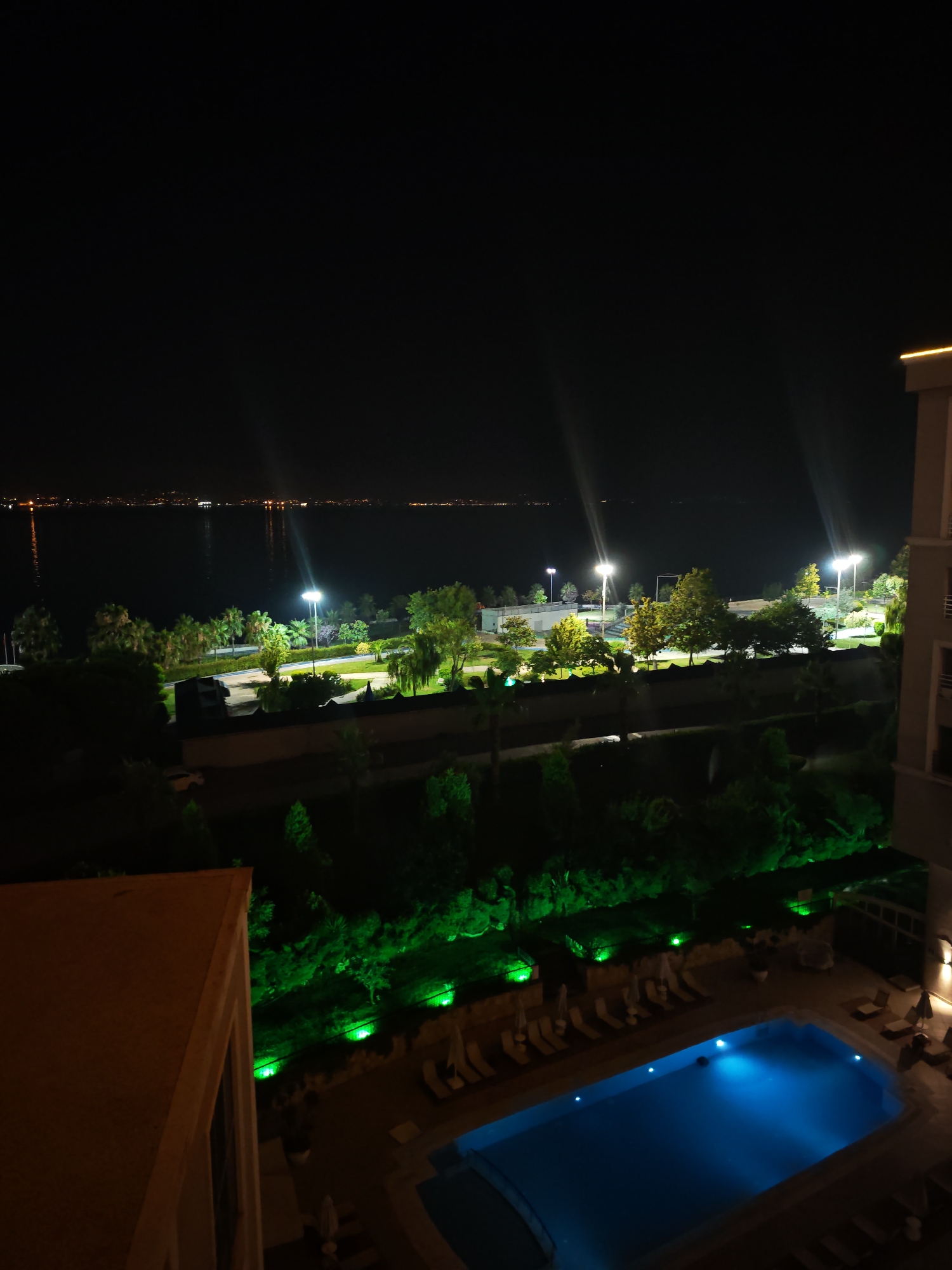The image is a nighttime aerial view of a resort or hotel in Darıca, Turkey, featuring a swimming pool illuminated by blue lights and surrounded by lounge chairs and umbrellas. Green lights highlight the lush vegetation and landscaping. In the background, a body of water, likely the Sea of Marmara, is visible with distant city lights reflecting on its surface. The scene depicts a serene and recreational environment, suggesting a holiday or leisure destination.