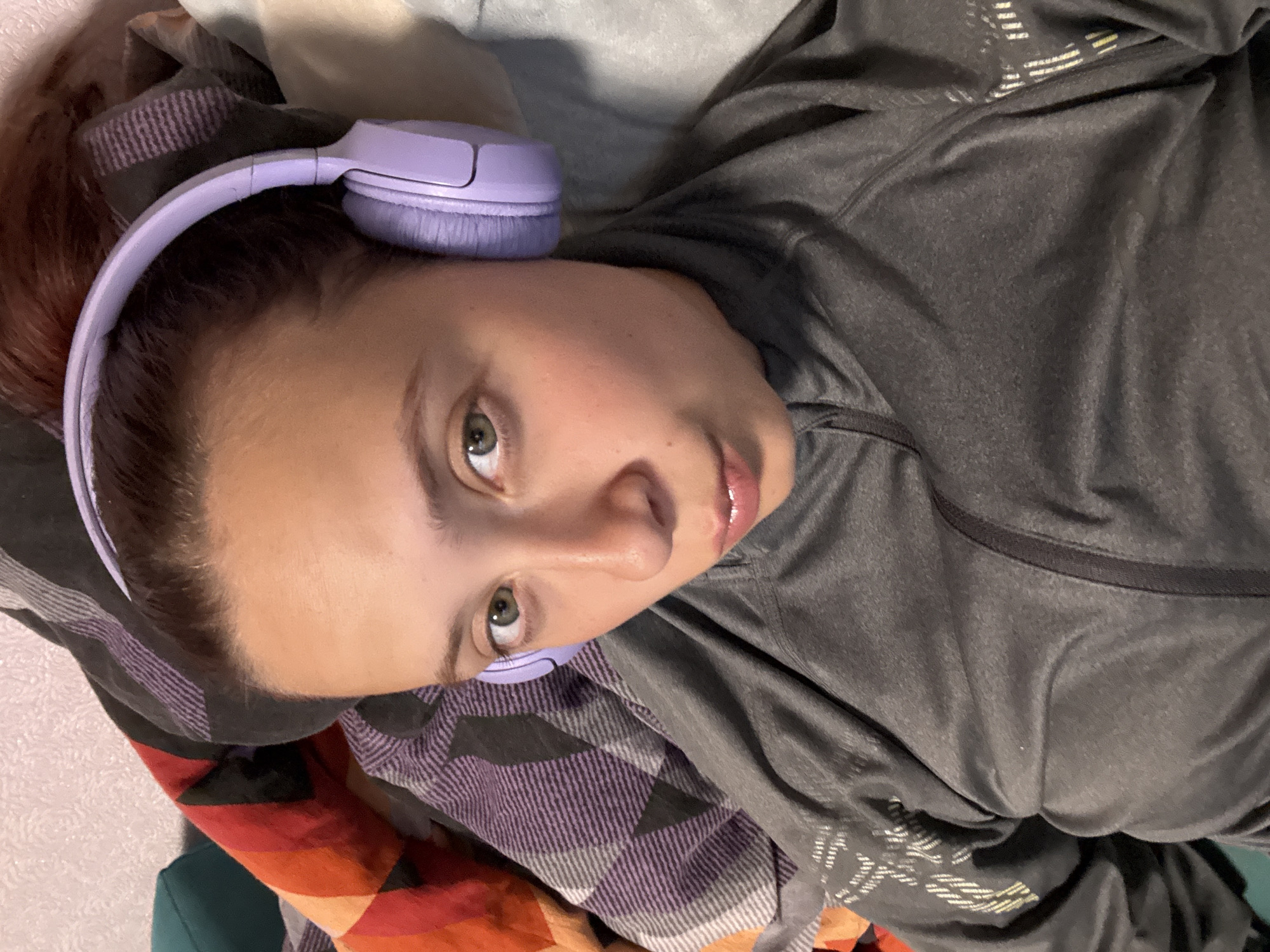 A young adult female, located in Прикамский, Russia, is depicted lying down indoors, looking directly at the camera. She has brown hair, pulled back, and green eyes, with a neutral expression and slightly parted, glossy lips. She is wearing light purple over-ear headphones and a dark gray long-sleeved athletic top featuring a partial front zipper and light-colored abstract linear designs on the sleeves. She is surrounded by textiles, including a multi-colored striped blanket (red, orange, black, purple) to her right, a purple, black, and white striped blanket behind her head, and a light gray textured blanket or pillow to her left. A light-colored, textured wall and a portion of a teal object are visible in the upper left background.