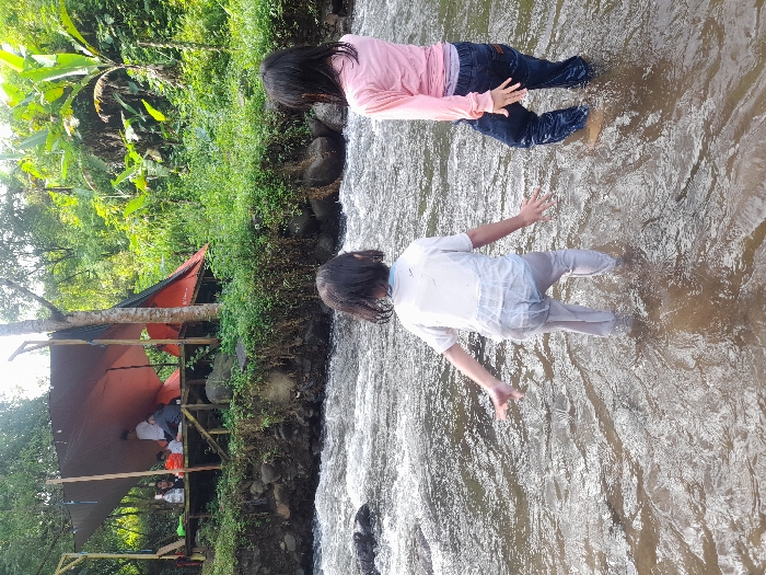 Two young people are wading in a shallow, flowing river. One wears a pink shirt and dark pants, while the other is in a white top and light pants. Both have their arms outstretched. On the riverbank, a makeshift shelter stands with people sitting inside. Greenery and trees surround the area, suggesting a natural setting. The water appears clear, and the sunlight creates bright reflections on the surface.