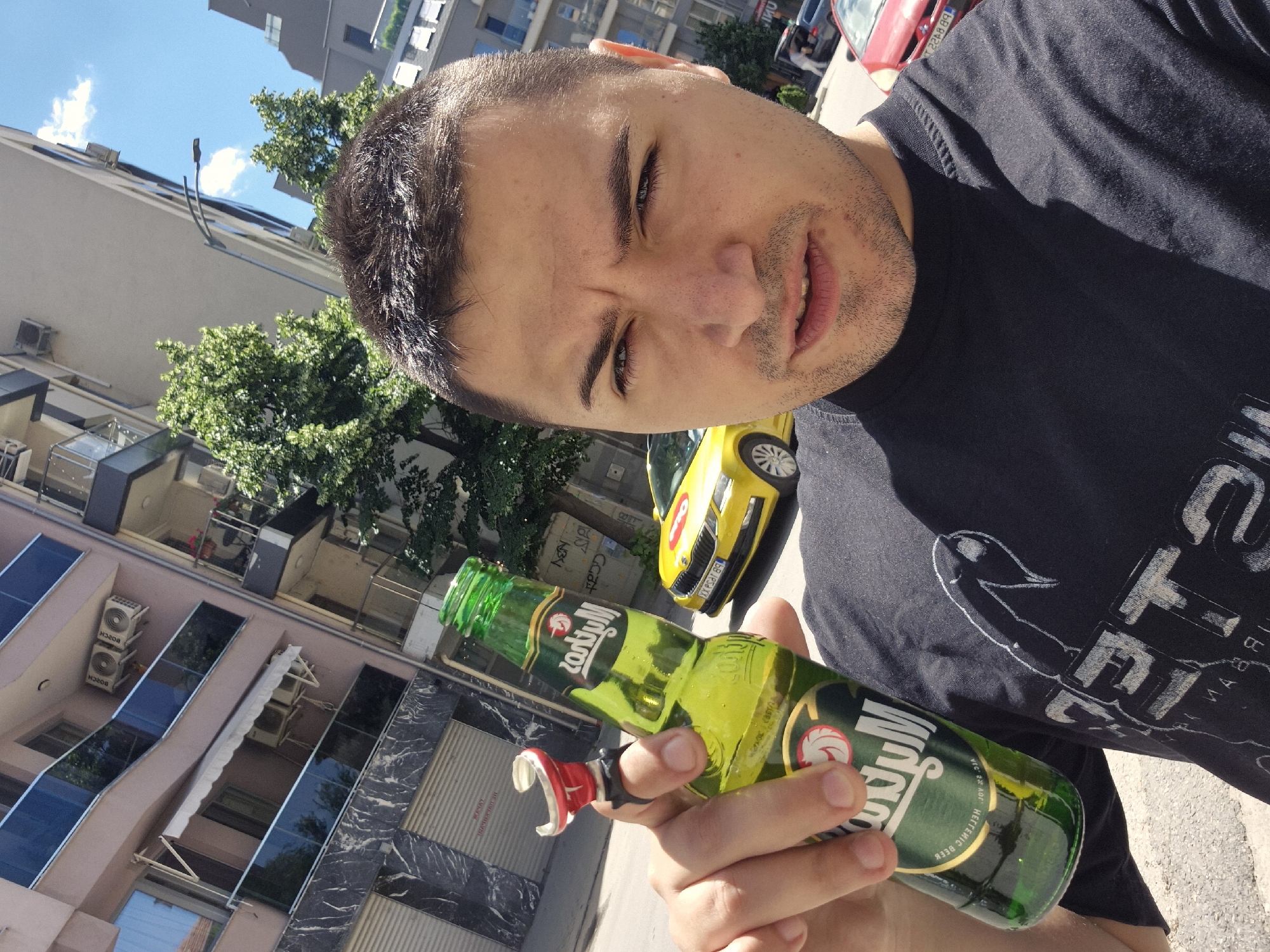 A man is taking a selfie while holding a bottle of Mythos Hellenic Beer. He is wearing a dark-colored t-shirt with a design on it. Behind him, there is a street scene with buildings, trees, and cars. A yellow car is parked close to the man. The buildings are light colored with balconies. The location appears to be Plovdiv, Bulgaria, based on the car's license plate and the architectural style.