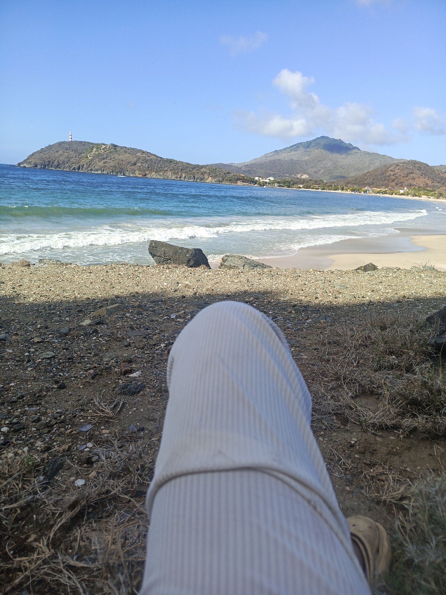 This is a first-person perspective image of a person sitting on a rocky, dry, grassy area overlooking a beach in Pedro González, Venezuela. The person is wearing light-colored, vertically striped pants and a brown shoe. The beach is a mix of sand and pebbles, with waves breaking on the shore. In the background, there are hills with sparse vegetation, and a lighthouse is visible on a distant hill. The sky is blue with some scattered clouds.