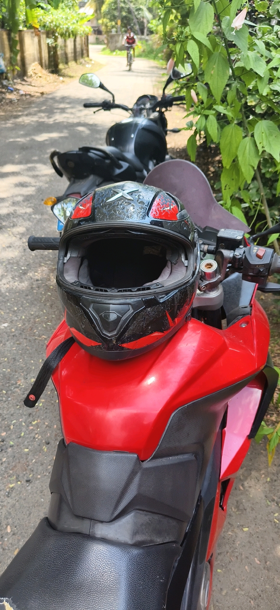 In Vaikom, India, a black and red motorcycle helmet rests on the front of a red motorcycle. The helmet has a textured black exterior with red accents and a silver "X" logo. A black strap is attached to the helmet, extending down and slightly to the left. Behind the red motorcycle, another black motorcycle is partially visible. The motorcycles are parked on a paved road lined with green foliage and a low stone wall. In the background, a person is seen riding a bicycle on the road. The scene appears to be outdoors, in a semi-urban or rural setting.