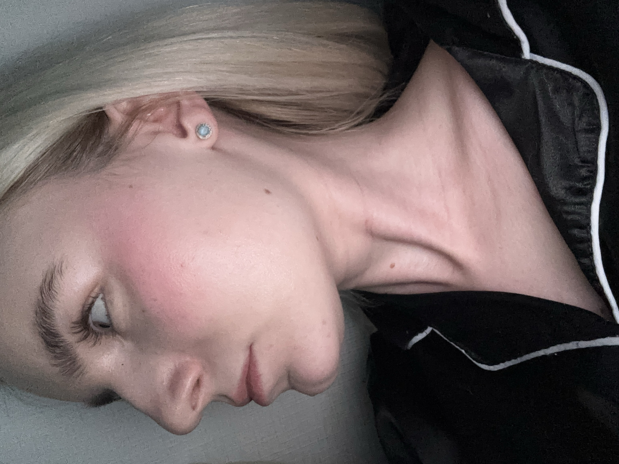 A close-up, side-profile image captures the head, neck, and upper shoulder of a person with light blonde hair. The individual's face is turned to the left, with the chin slightly tucked. Their fair skin is evident, highlighted by a flush of pink blush on the cheek. A small, light blue stud earring is visible in the left earlobe. The left eye, with its dark and prominent eyebrow and long eyelashes, is partially visible, gazing upwards or to the side.
The neck area features visible veins and tendons, suggesting a specific posture or moment of slight tension. A black garment with distinctive white piping, possibly a pajama top or a satin robe, covers the right shoulder and chest area. The hair, styled relatively straight, flows over the ear and partially obscures the top of the head.
The background is softly blurred, appearing as a light grey surface behind the head and a darker, undefined soft surface beneath the chin and neck, reinforcing an indoor setting. The lighting is soft and even, highlighting the contours of the face and neck. There is no visible text in the image. The overall impression is an intimate, perhaps artistic, portrait focusing on the details of the subject's profile and skin texture.