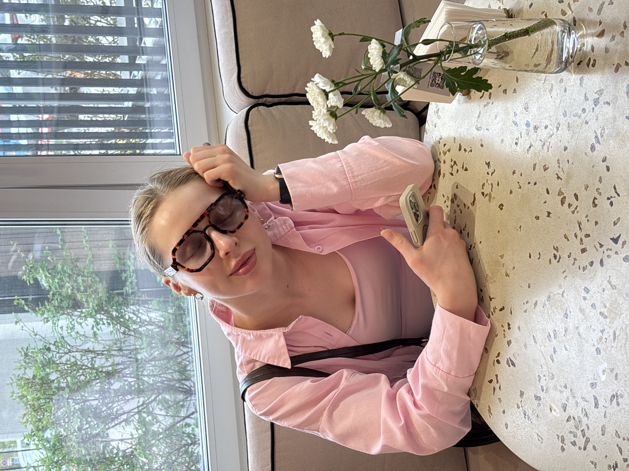 A woman wearing glasses and a pink shirt sits at a table in a cafe in Perm, Russia, holding a smartphone. The cafe has a window overlooking foliage and a flower arrangement in a glass vase on the table. The table has a speckled surface. The woman is resting her head on her hand.