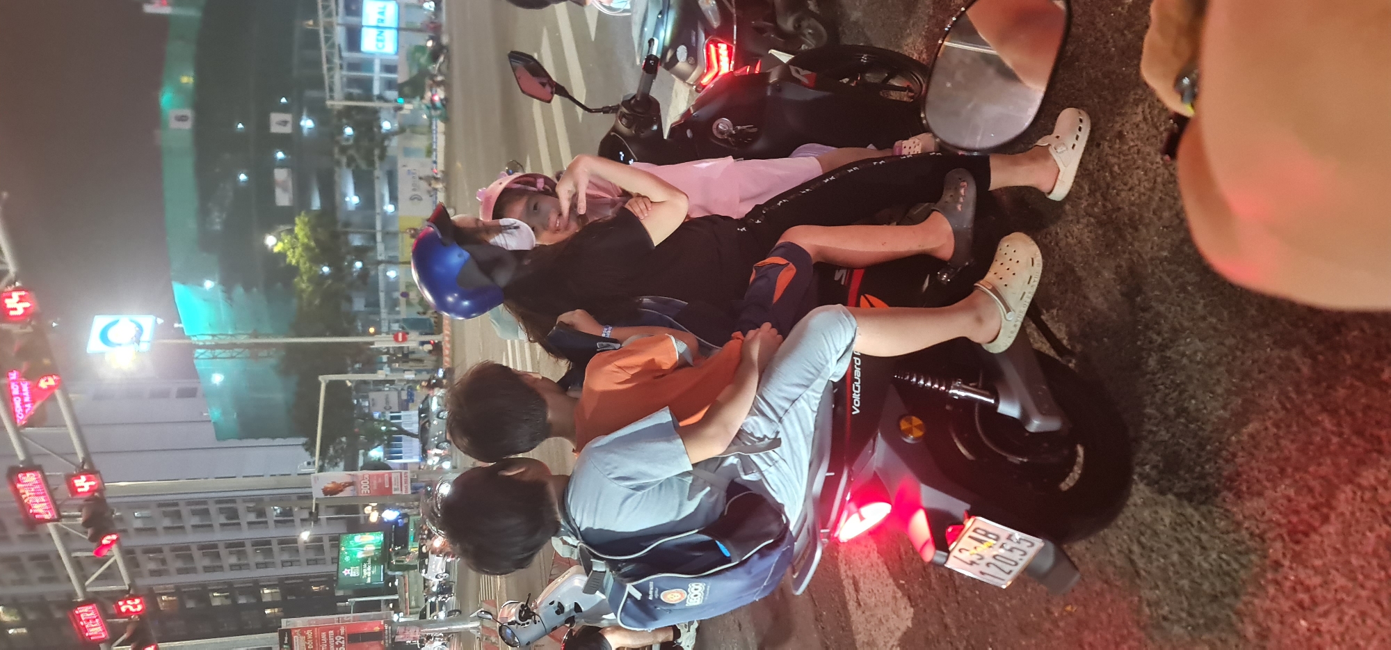 The photograph shows a family of four people sitting on two motorbikes on a city street. The location is identified by license plates and building signs to be in the city of Đà Nẵng, Vietnam. The children, a young boy in a blue helmet, and a young girl with a pink headband are on the front bike. The driver is obscured and the back driver is only partially visible. Buildings are visible in the background with signs in Vietnamese and English. At least one of the motorbikes is manufactured by VoltGuard. The license plate number is 43-AB 120.55.