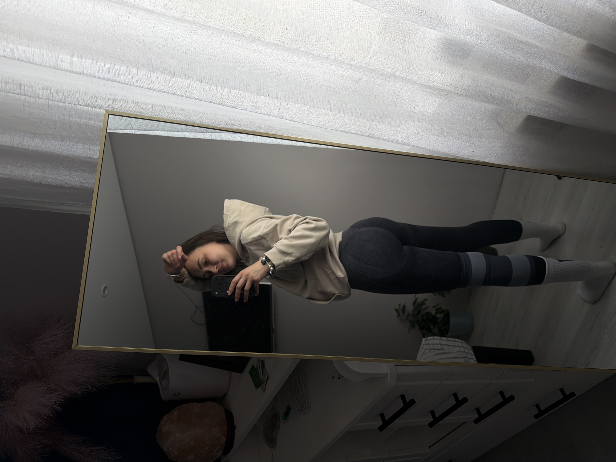 A woman is reflected in a full-length mirror. She is wearing a beige hoodie and dark-colored leggings with striped sections at the lower legs. She holds a black smartphone in her right hand, presumably taking the photograph. The background reveals a room with a white curtain, a white dresser with black handles, a salt lamp, and a potted plant. The reflection also captures a framed picture on the dresser. While the location is suspected to be Chaikovsky, Russia, no elements in the photo explicitly confirm this.