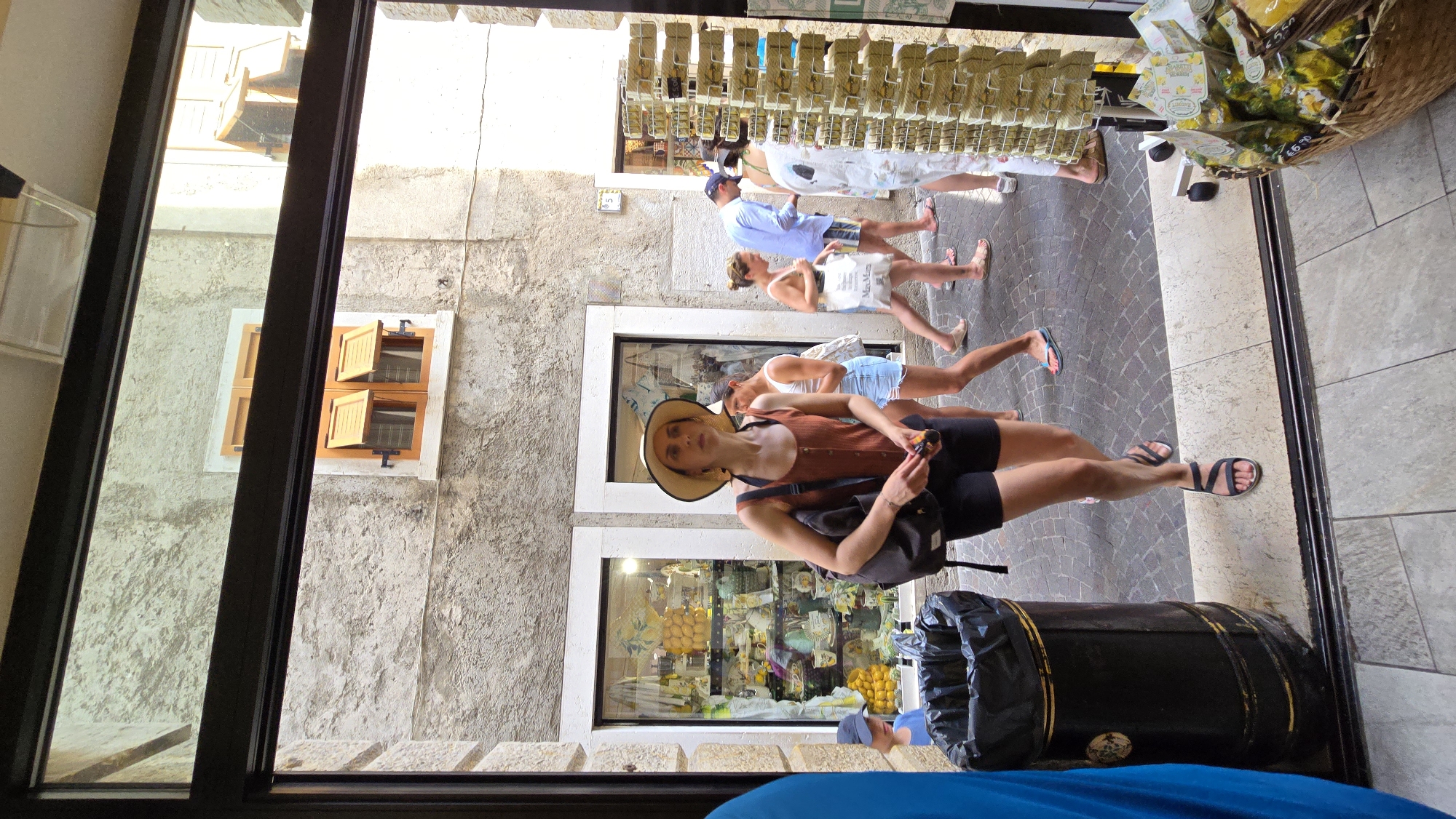 This image captures a street scene in Limone sul Garda, Italy, looking into a shop or arcade. In the foreground, a woman wearing a wide-brimmed hat, a rust-colored tank top, and black shorts stands facing forward, holding a small dark object in her right hand. She has a dark shoulder bag slung across her body. To her left is a large, dark trash can with yellow trim. Behind her, several other people are visible. To her right, a woman in a white tank top and denim shorts walks away. Further back, a man in a blue shirt and patterned shorts walks past. The buildings have stone facades and wooden shutters on the windows. Display shelves filled with goods are visible in the shop windows and an outdoor display rack is to the right. The ground is paved with cobblestones.