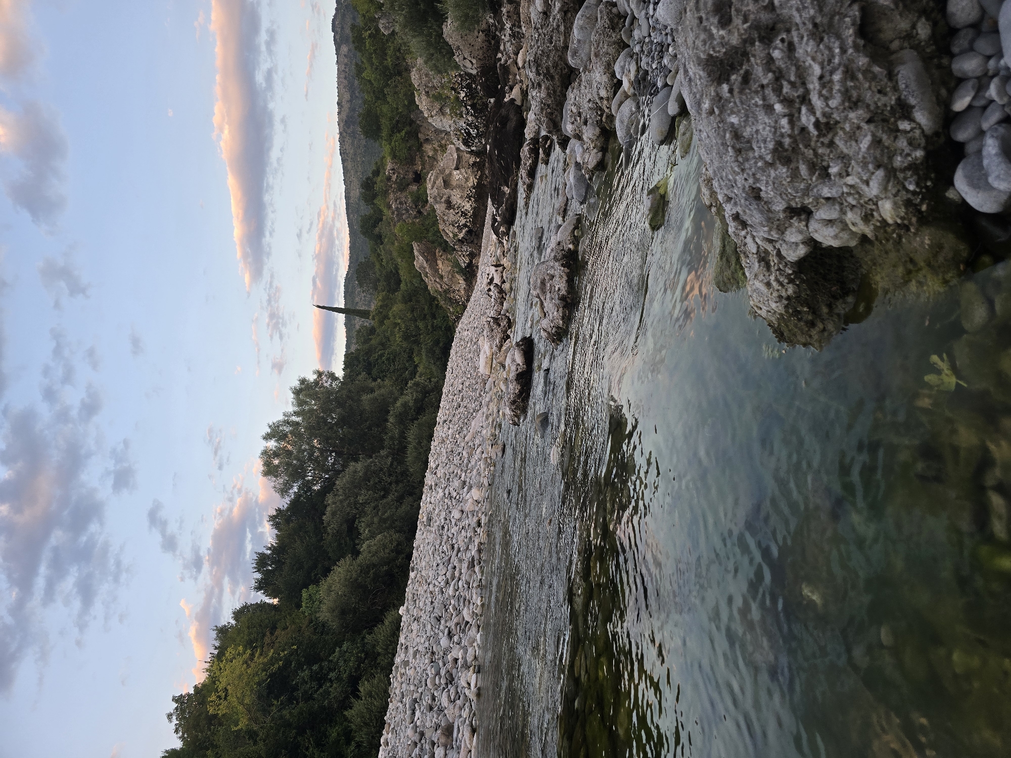 The image depicts a shallow, clear river flowing through a rocky terrain. The riverbed is composed of smooth, grey stones of various sizes. The water is transparent, allowing the submerged stones and hints of green aquatic vegetation to be visible. The banks of the river are lined with more of these grey stones, forming a gravelly shore. Beyond the river, a lush green hillside rises, densely covered with trees and shrubs. In the distance, hills are visible, with a tall, slender tree standing out against the skyline. The sky above is a soft blue, with scattered clouds tinged with pink and orange from what appears to be sunset or sunrise. The overall scene is natural and serene, suggesting a remote, unspoiled environment. The location is specified as Bioče, Montenegro.