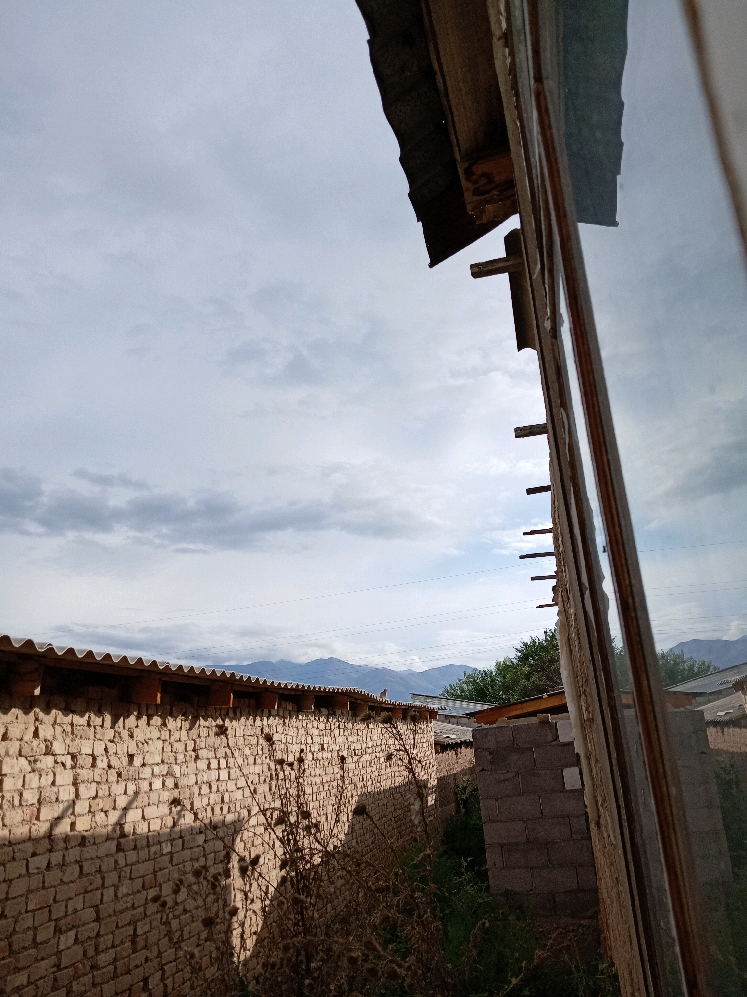 The image shows a view from what appears to be a window frame, looking out onto a brick wall and a cloudy sky. The brick wall is made of light-colored bricks and is topped with a corrugated metal roof. To the right of the brick wall, there is another structure made of concrete blocks. In the distance, mountains can be seen under a cloudy sky. The overall scene suggests an outdoor environment, possibly in a rural or semi-rural area. The presence of the window frame suggests the photograph was taken from inside a building. The location is identified as Chaldybar, Kyrgyzstan.