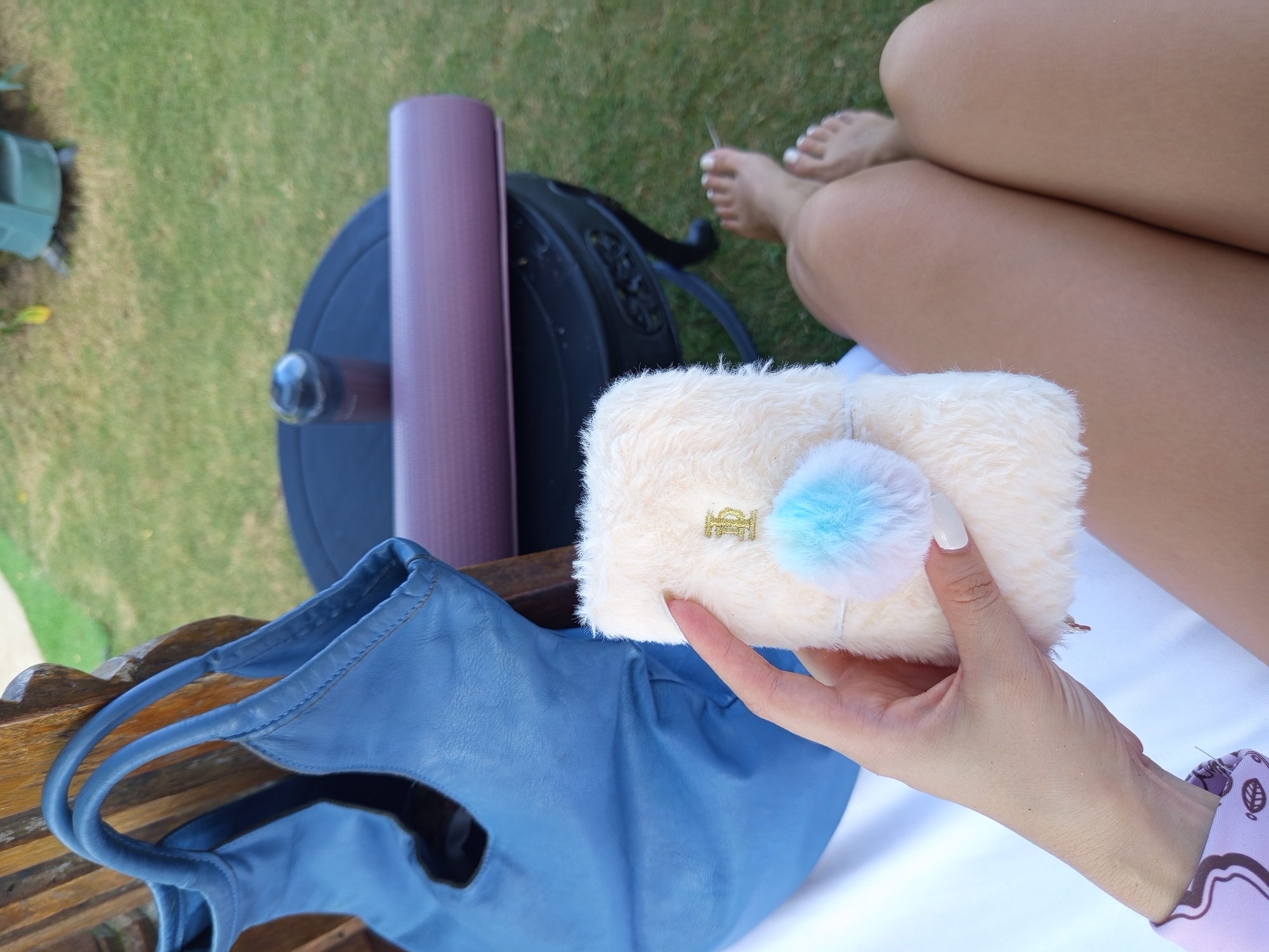 This overhead shot captures a serene outdoor leisure scene in North Kuta, Indonesia. The primary subject is a person relaxing, with their tan legs, featuring white-painted toenails, visible on the right side of the frame, resting on a white surface. A hand, also appearing from the right, holds a fluffy, cream-colored pouch. This pouch is embellished with a small, golden, ornate emblem, possibly a stylized "BB," and a blue and white pom-pom.
In the midground, a blue, soft-textured tote bag sits on a wooden surface, likely a bench or chair. Behind the bag, a rolled-up purple yoga mat is positioned next to a dark, somewhat circular object that appears to be a backpack or another bag. The background is dominated by lush green grass, suggesting a well-maintained garden or lawn area. A small, blurry green plastic container is partially visible on the far left. The natural lighting indicates it is daytime, contributing to the tranquil atmosphere. The arrangement of the yoga mat and bags suggests the person might be enjoying a break before or after a fitness activity, or simply relaxing during a tropical getaway. No clear, readable text in any language is visible, only the stylized emblem on the pouch.