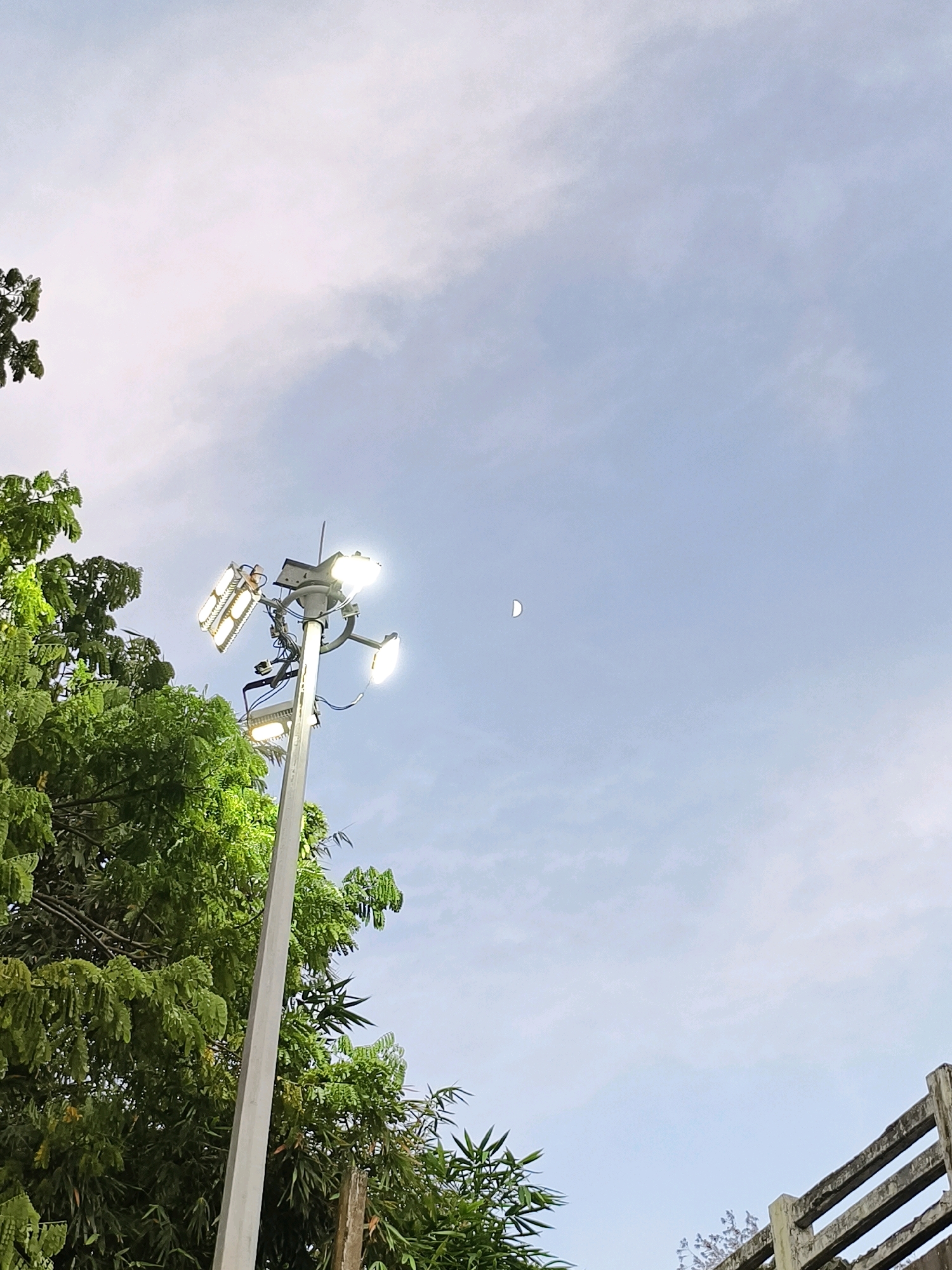 The image captures a daytime scene with a clear blue sky and scattered clouds. In the foreground, a tall, grey lamp post is visible, equipped with multiple bright LED lights, some of which are illuminated. Lush green foliage from trees frames the left side of the lamp post, with bamboo-like plants also present. In the upper right quadrant of the sky, a sliver of the crescent moon is discernible. In the lower right corner, a concrete structure resembling a fence or railing with horizontal beams is present. The scene appears to be outdoors, potentially in a park or public area, given the presence of street lighting and surrounding greenery. The location is specified as Anangadi, India.