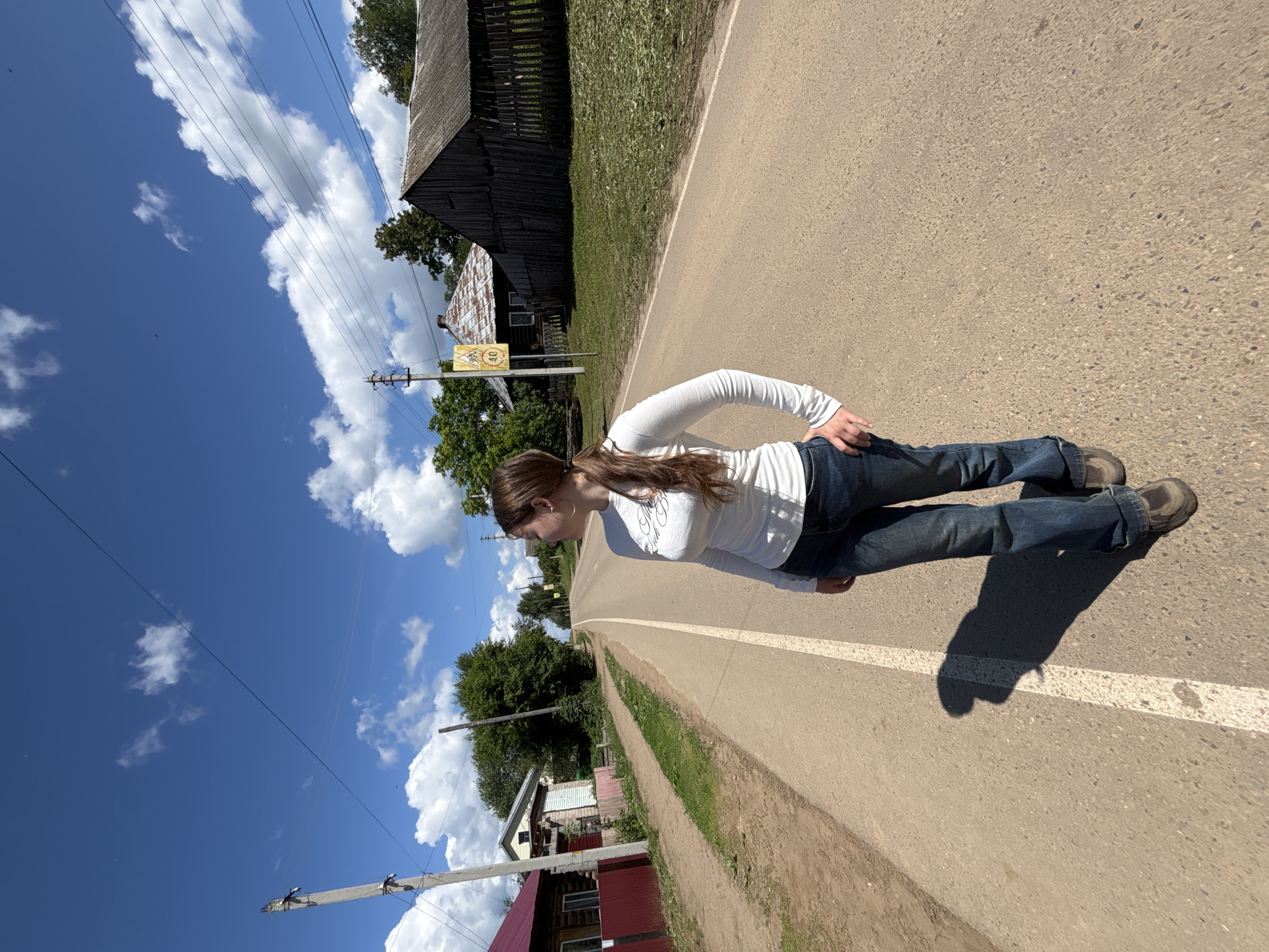 The scene is depicted in Фоки, Russia, on an asphalt road under a bright blue sky with scattered white cumulus clouds. A young adult female is positioned on the road, slightly right of the frame's center. She possesses long, light brown hair secured in a ponytail and is dressed in a white long-sleeved top, blue denim jeans, and dark brown or grey athletic shoes. Her right hand rests on her hip or in her pocket. She is oriented slightly to her left, with her head turned to the right, looking horizontally away from the camera. A distinct shadow of the female is cast upon the asphalt road to her left.
The asphalt road features a faded white dividing or edge line. To the left of the road, a reddish-brown dirt shoulder is present, bordered by a red-painted wooden picket fence. Behind the fence, a log cabin-style house with a red roof and white window trim is partially visible. Further back, another house with a light-colored facade and dark roof is discernable. A concrete utility pole with attached power lines stands vertically on the left side of the frame.
On the right side of the road, a yellow diamond-shaped road sign with a black border, depicting two children, is present. Below it, a rectangular sign indicates "40" in a red circle, denoting a speed limit. Beyond this sign, a weathered, dark wooden structure with a corrugated metal roof and a wooden fence is situated. Green foliage and trees line both sides of the road. Multiple dark power lines traverse the upper portion of the blue sky, with small dark specks, possibly birds, visible against the sky.