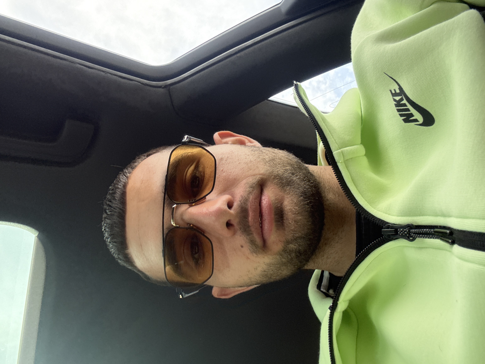 A man wearing sunglasses and a light green Nike zip-up sweatshirt is inside a vehicle with a sunroof. The location is indiscernible and there is no information available within the image to suggest the location is Pirgovo, Bulgaria. The camera angle is from above, looking down at the man.
