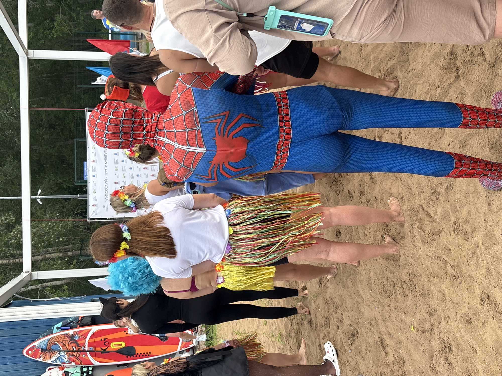 In Chaikovsky, Russia, a group of people are gathered on a sandy beach near a wooden structure. The central figure is dressed in a full Spiderman costume, facing away from the camera. Several other individuals are visible, some wearing Hawaiian-themed attire, including leis and grass skirts. One person has a blue wig, and another wears cat ears. A paddleboard with orange and white accents is propped up on the left side of the frame. The background shows trees and a white banner with Russian text. The scene appears to be a casual outdoor event or gathering.