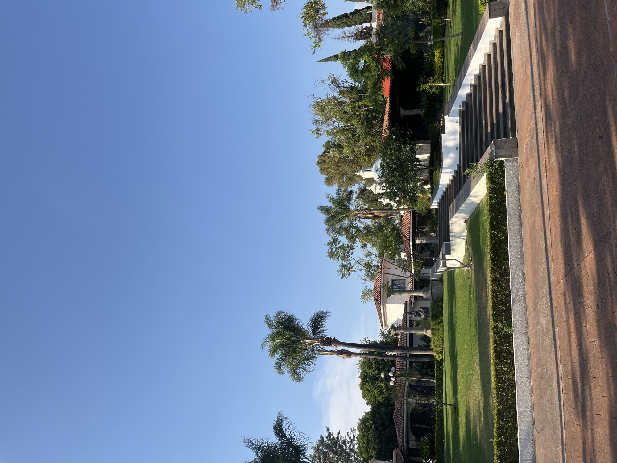 The image depicts a partially sunny day scene in San Bernardino Tlaxcalancingo, Mexico, showcasing a landscaped area. The scene is dominated by a clear blue sky. The right side of the image reveals a staircase structure with dark steps and white risers, adjacent to a brick walkway. A green lawn is visible, bordered by trimmed hedges, leading to a white building with a red tiled roof. Several palm trees and other trees are present, contributing to the landscape.