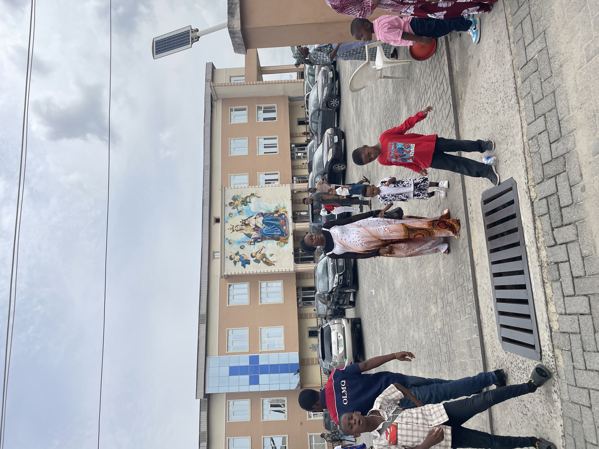 Here is a detailed description of the image:
The image shows a group of people standing in front of a multi-story building in an urban environment, likely in the city of Unknown, Unknown. The building has a beige facade with multiple windows and a large mural depicting a religious scene with figures that appear to be angels and a central female figure. There's also a section of the building with blue and white checkered glass panels.
Several cars are parked along the side of the building. The sky is overcast with a greyish-white hue, indicating a cloudy day. A solar-powered street lamp is visible near the top of the frame, suggesting this area is striving for sustainability.
The people are of various ages, including children and adults. One child wears a red shirt with a Spider-Man graphic, another has a black-and-white patterned outfit, and an adult woman is wearing a white and orange patterned garment. Some of the people are looking towards the camera.
The ground is paved with grey bricks and has a drainage grate visible. The environment appears to be a public space, possibly a gathering area or a parking lot. The presence of power lines is noticeable against the sky.