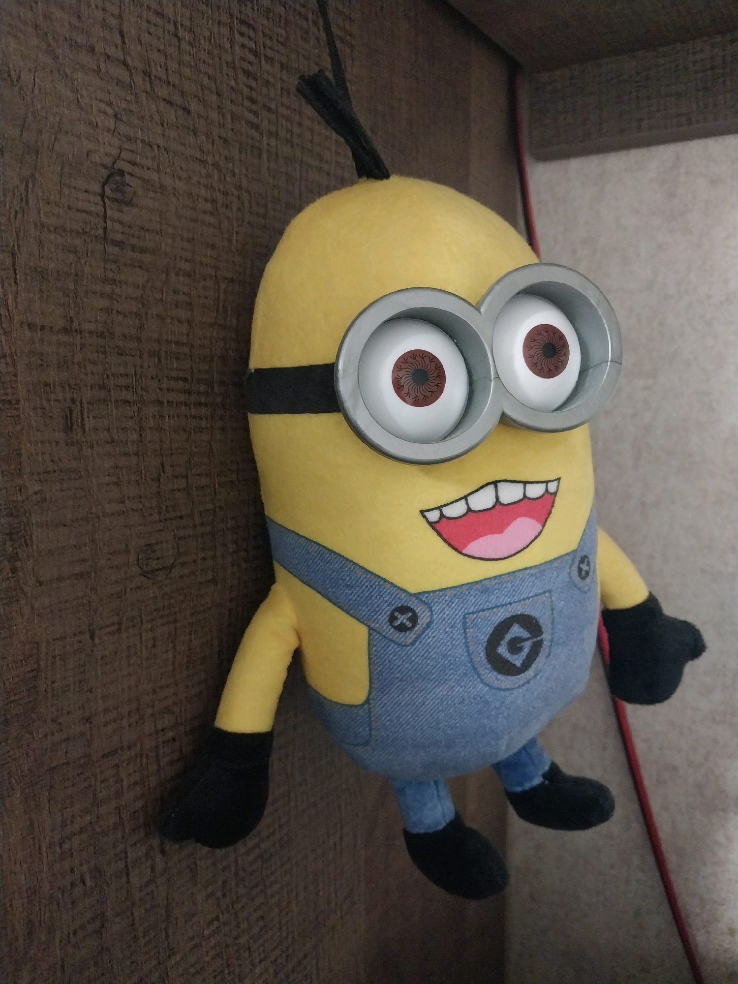A plush toy of a Minion character from the Despicable Me franchise is positioned on a wooden surface. The toy is yellow with large goggles and is wearing blue overalls. The background appears to be a wall with a textured surface. The image is a photograph taken in Dnipro, Ukraine.