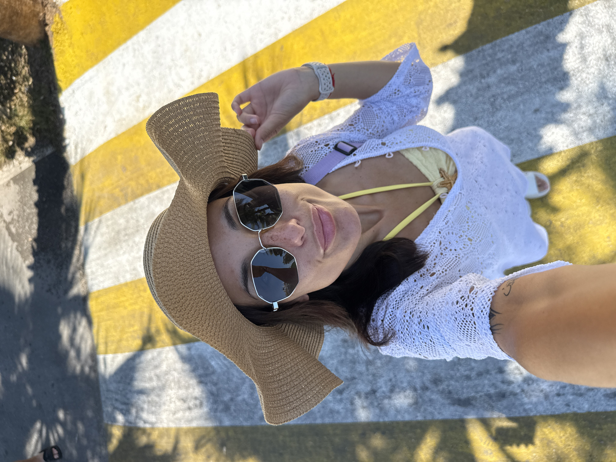 In the city of Unknown, Türkiye, a woman is taking a selfie while standing on a pedestrian crossing with yellow and white stripes. She is wearing a wide-brimmed straw hat, sunglasses, and a white crocheted cover-up over a yellow bikini top. She has freckles on her face and a tattoo on her left arm. A purple strap from a bag is visible over her shoulder. The sun casts shadows on the ground.
