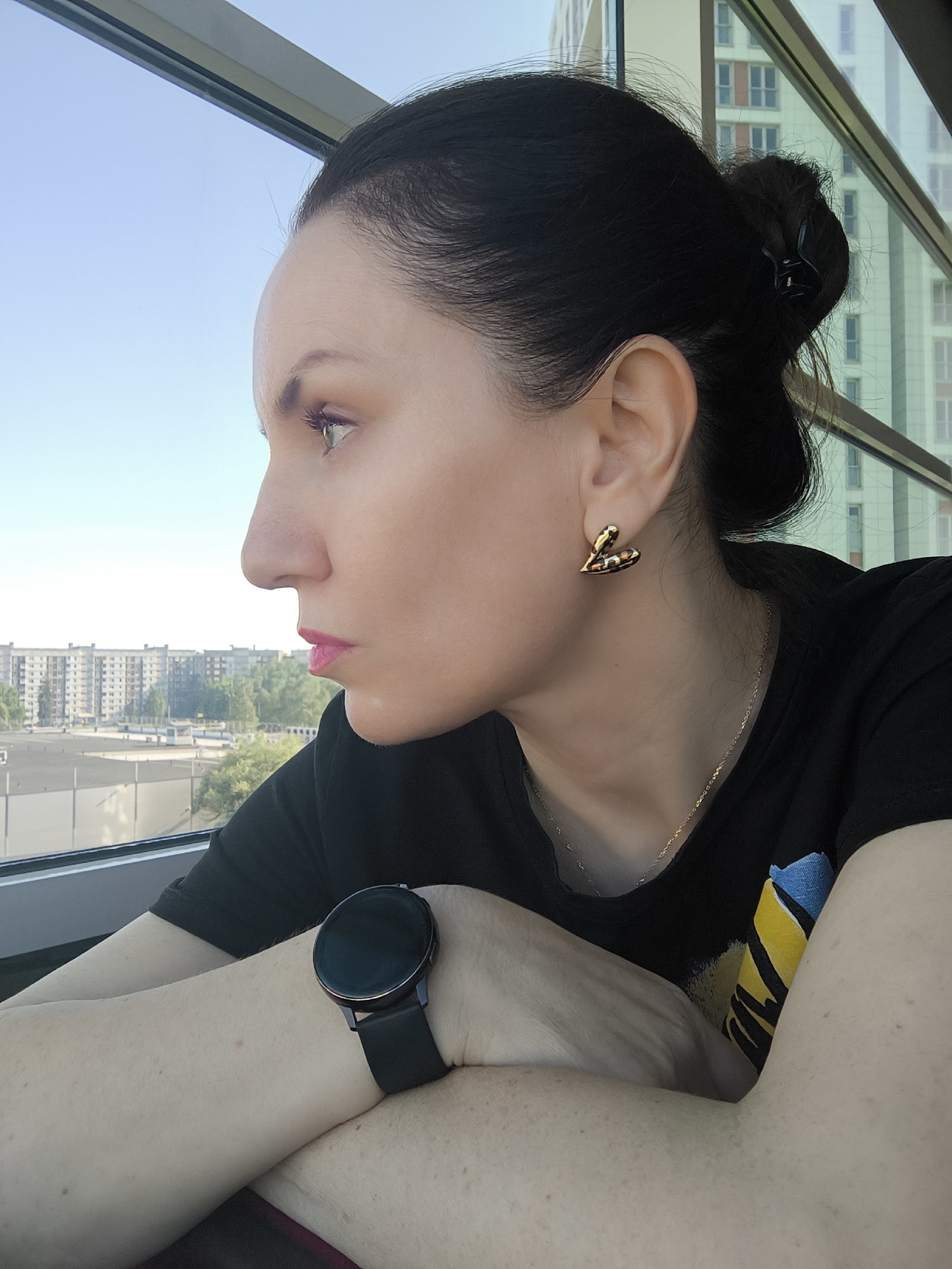 A woman is depicted in a profile shot, looking out a large window at an urban landscape during the daytime. Her dark hair is pulled back in a bun, secured with a small black claw clip. She wears heart-shaped, textured gold-toned earrings and a thin gold chain necklace. Her lips are adorned with bright pink lipstick.
She is dressed in a black t-shirt, part of which shows a blue and yellow design on the front. Her arms are folded or resting on the windowsill, with a black smartwatch visible on her left wrist. The watch has a round face and a dark strap. Her skin tone is fair, with visible freckles or moles on her arms.
Beyond the window, a bright, clear blue sky is visible. The outdoor scene features several multi-story residential or office buildings, some appearing older and blocky, others more modern with numerous windows. Green trees are interspersed among the buildings, suggesting an urban or suburban setting. The overall mood is contemplative or observant as she gazes out. There is no visible text in the image.