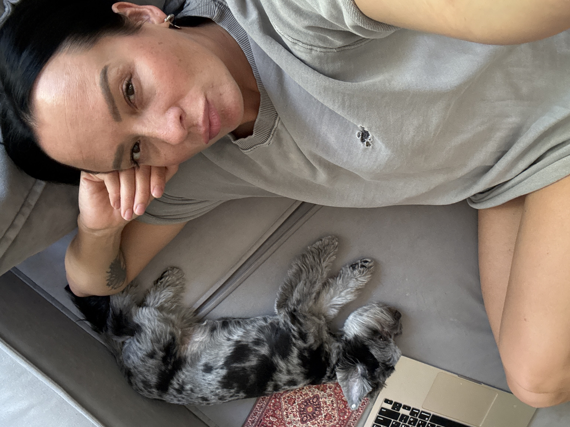 A woman is lying on a gray couch next to a small dog. The woman has dark hair and is wearing a gray sweatshirt. She is resting her head on her hand and looking at the camera. The dog is gray and black with a mottled coat. It is lying on its side, facing away from the camera. A laptop is on the couch to the left of the dog. A small rug with a red and gold pattern is partially visible next to the dog. The overall lighting is natural and appears to be daylight. The image does not contain any information that places it in the city of Chaikovsky, Russia.