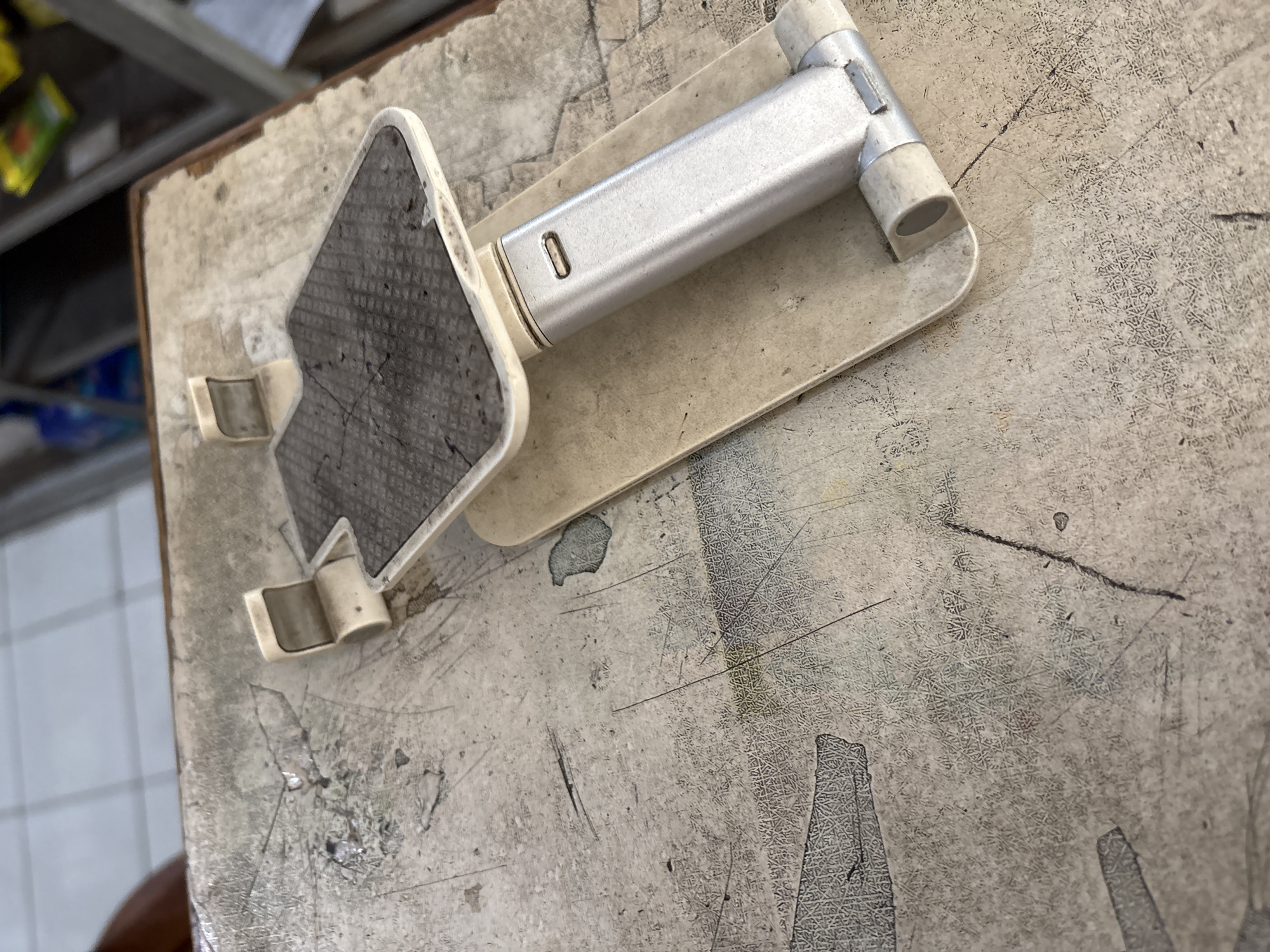 The image shows a close-up, eye-level view of a portable phone stand on a worn, textured surface. The phone stand is primarily made of light-colored plastic and brushed aluminum, with a dark grey textured grip pad on the top platform. The surface beneath the stand appears to be a table or countertop with a distressed, mottled finish, exhibiting numerous scratches, marks, and some darker stains. The background is blurred but suggests an indoor setting, possibly a shop or storage area with shelves and various items visible. The lighting is even, casting soft shadows. There are no people or animals in the image. This scene is presented without any specific context or indication of its location in Palimanan, Indonesia.