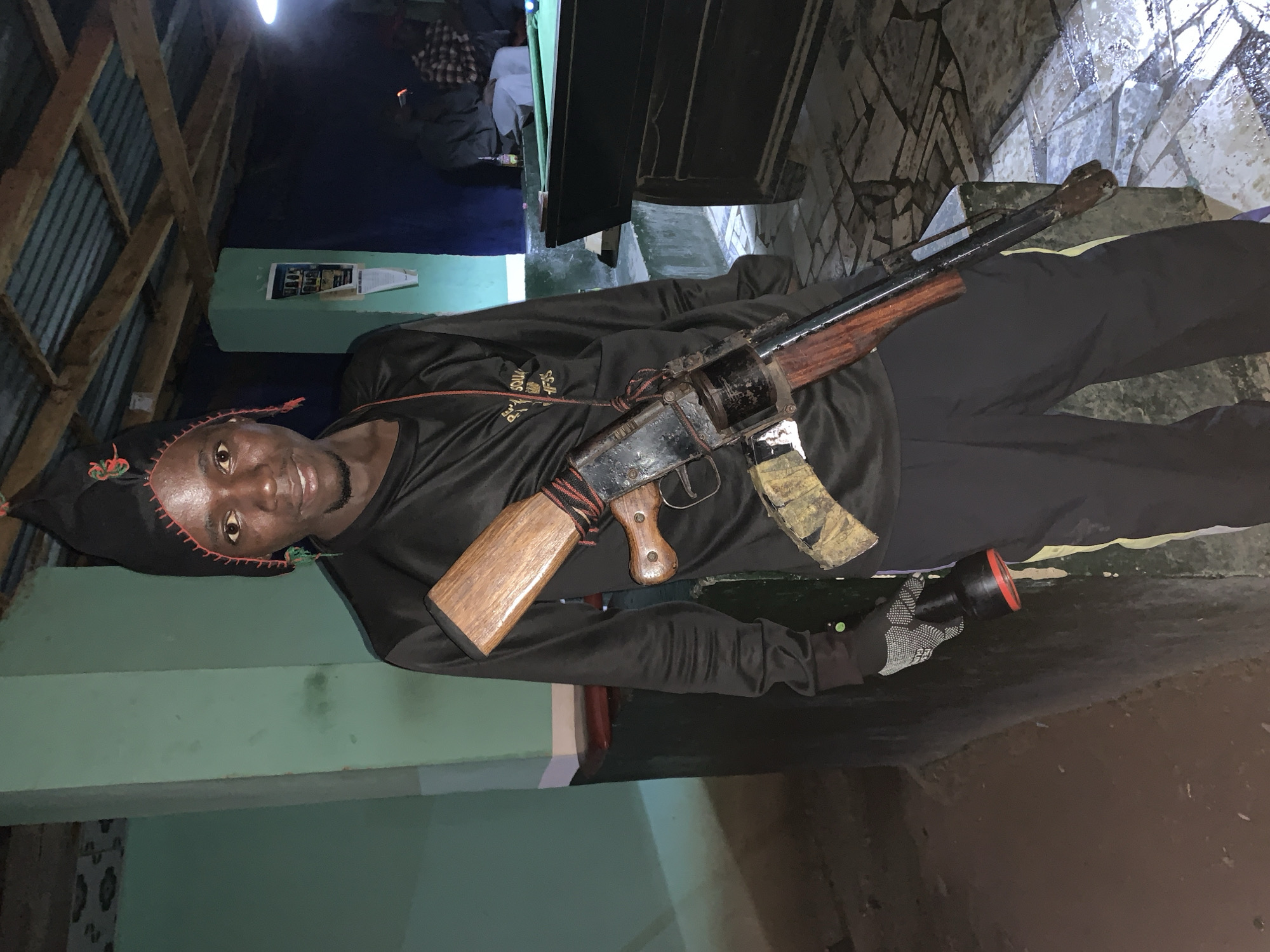A man wearing a black skull cap and black clothing poses holding a homemade firearm and a flashlight. He is standing indoors with walls of different colors, potentially green and white. The firearm is predominantly black and brown, and it has a magazine inserted. The setting is a building in New Stadium, Nigeria.