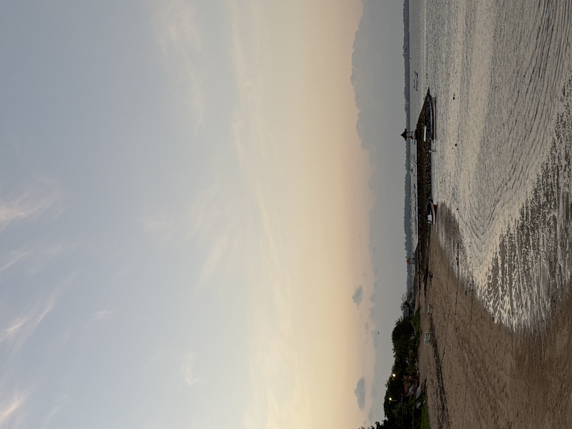 A tranquil beach scene unfolds in Kuta Selatan, Indonesia, as the sky transitions through hues of pale blue and soft orange, indicating either dawn or dusk. The sandy shore, marked by the receding tide, reveals intricate patterns of wet sand. A stone jetty juts into the calm, grey water, supporting a small, dark pavilion. Several boats are moored around the jetty, with one white boat with a red stripe visible closest to the shore. Lush green trees and vegetation line the edge of the beach in the background, interspersed with small structures. The overall atmosphere is serene and quiet, characteristic of a peaceful coastal environment.