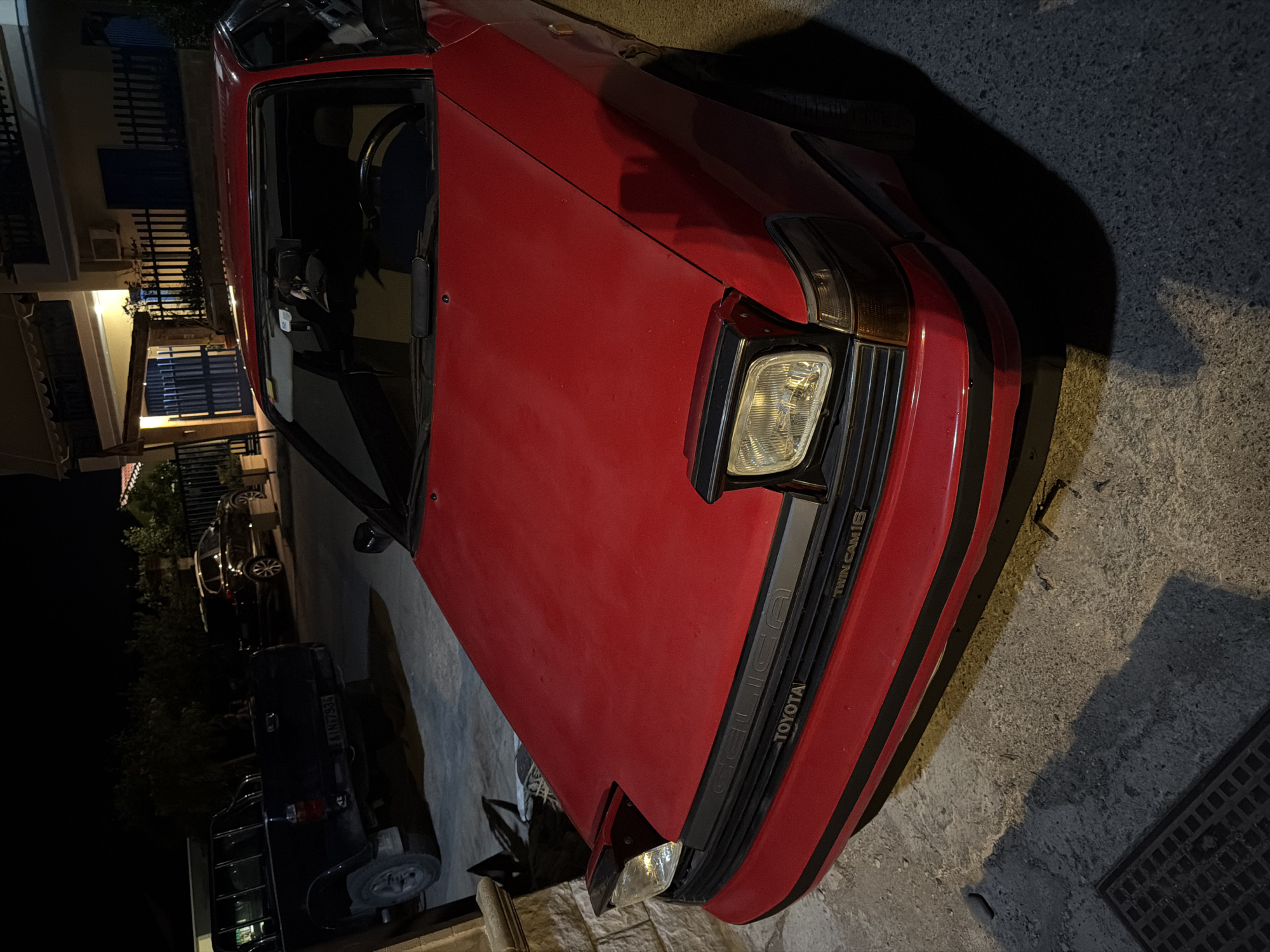 An aged red Toyota Celica GT-S with pop-up headlights is parked on a paved surface at night in front of a residential building. The car is positioned at an angle, with its front prominently displayed. The pop-up headlights are partially raised, with one illuminating. The car features a black grille with "TOYOTA" and "TWIN CAM 16" badging. A black pickup truck is parked to the left of the Celica. The background shows a building with a balcony and a dark fence. The scene appears to be in the Municipal Unit of Alyzia, Greece, indicated by the license plate format on the pickup truck. The lighting suggests it is nighttime.