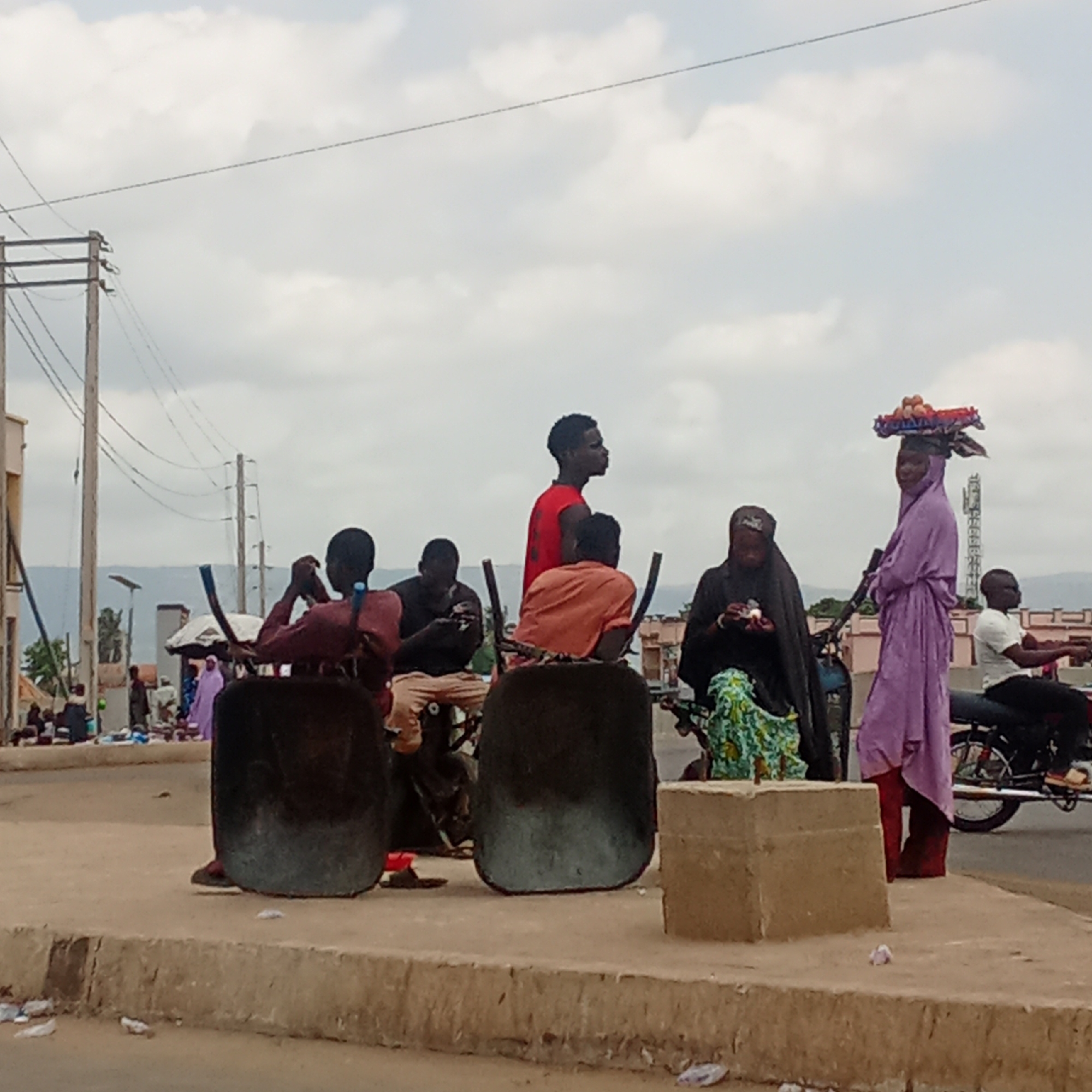 In Akwanga, Nigeria, a group of men is seated on stools with high backs along a concrete curb at the edge of a road. A woman wearing a black hijab sits behind them on a motorcycle, a woman wearing a purple hijab and carrying a tray of goods on her head standing near her. A man on a motorcycle is parked near the woman in purple. Overhead power lines and a telecommunications tower are visible in the background. Litter is visible on the road.
