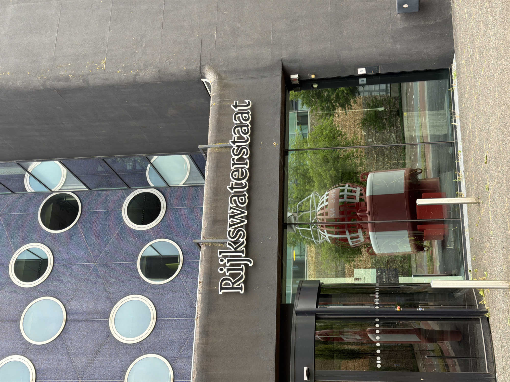 The exterior of a modern building is shown, featuring a dark grey facade on the right and a patterned blue facade with circular windows on the left. A prominent sign above a glass entrance reads "Rijkswaterstaat". Inside the glass-fronted area, a large, red and white buoy-like sculpture is visible. A revolving glass door is on the left side of the entrance. The scene appears to be in a city, with trees and a road visible through the glass. The location is Rijswijk, Netherlands.